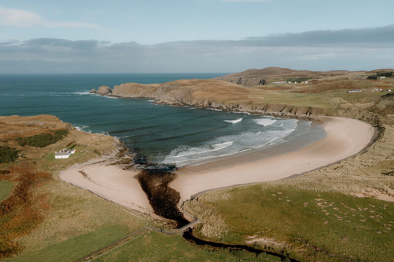 A curved sandy bay framed by headlands and grassy hills, seen from above.