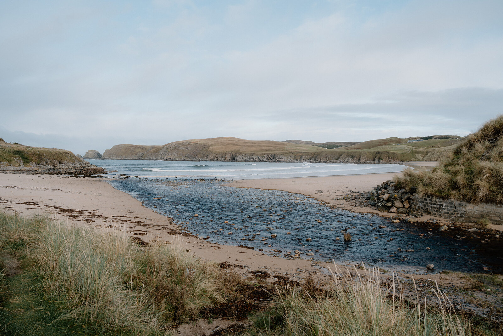 A tidal inlet winding through dunes to meet the sea, distant sea stacks visible on the horizon