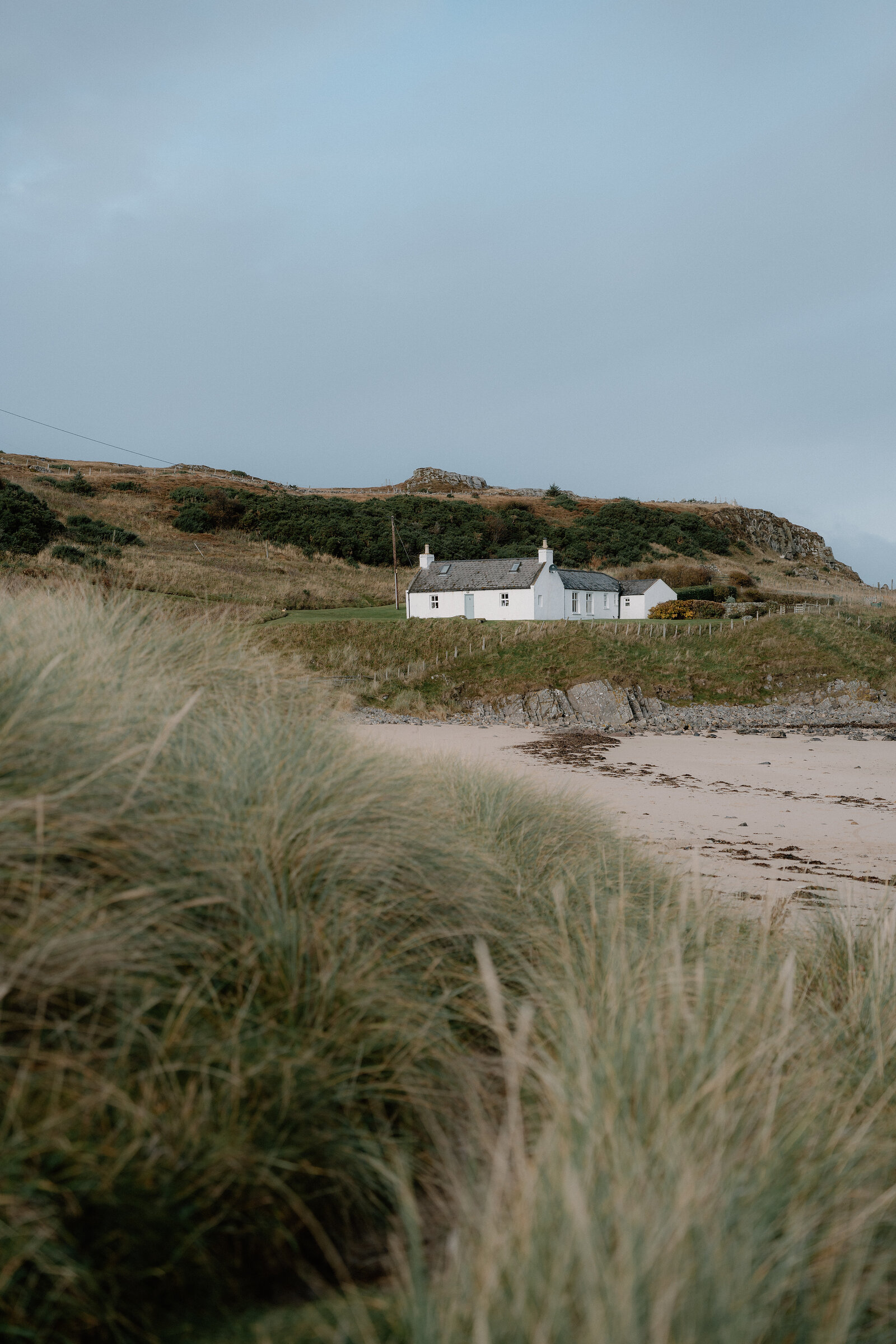 A white cottage set above a curved sandy beach, dune grass in the foreground