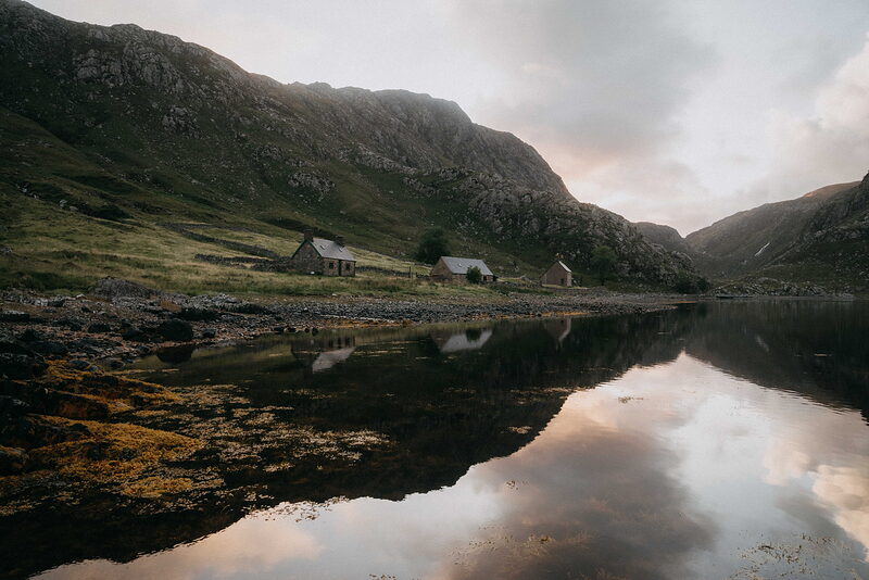A row of stone cottages beside a still loch, mountains rising steeply behind and reflected in the water.