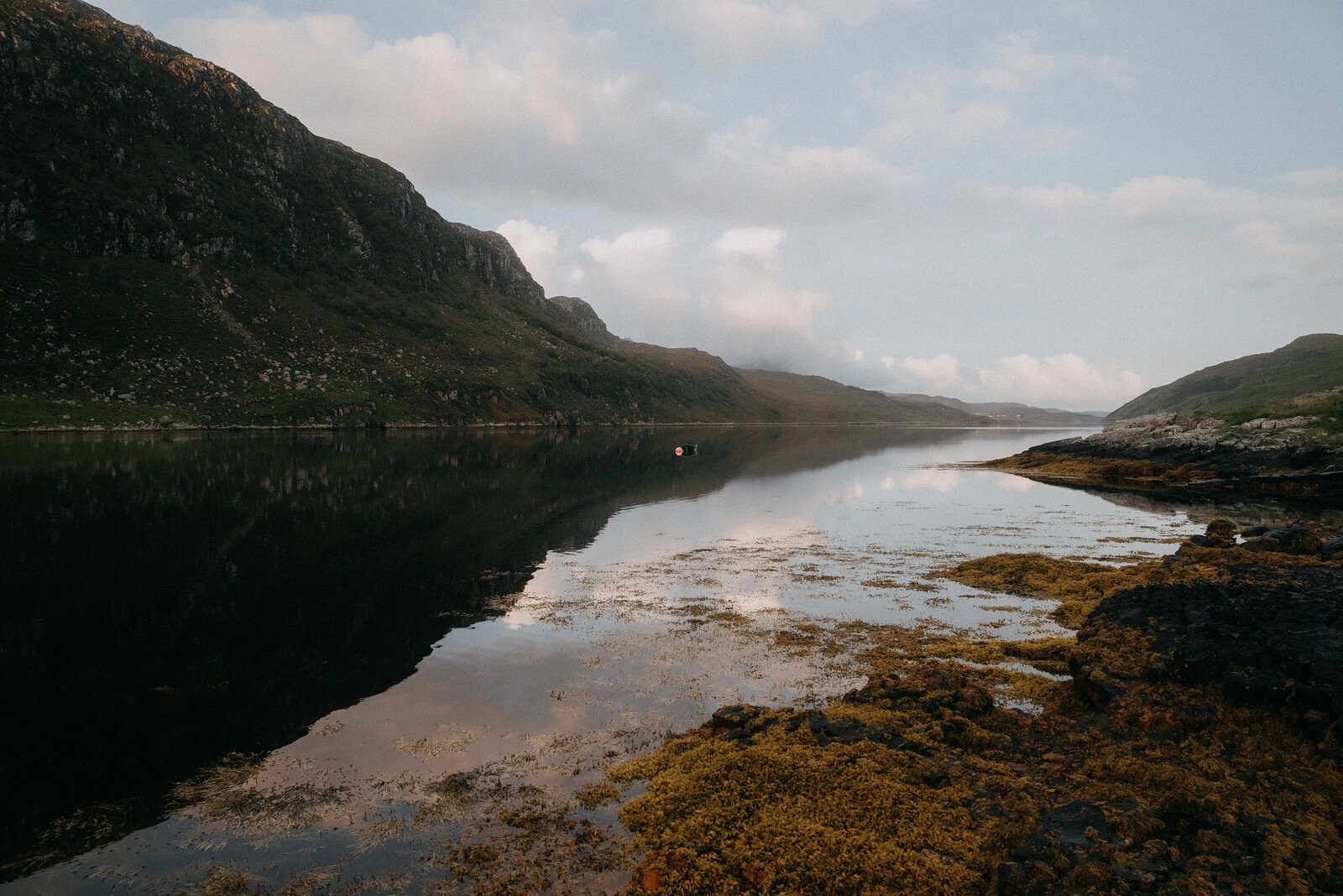 A still loch mirroring steep mountains under a pale sky, seaweed-covered rocks along the shore.
