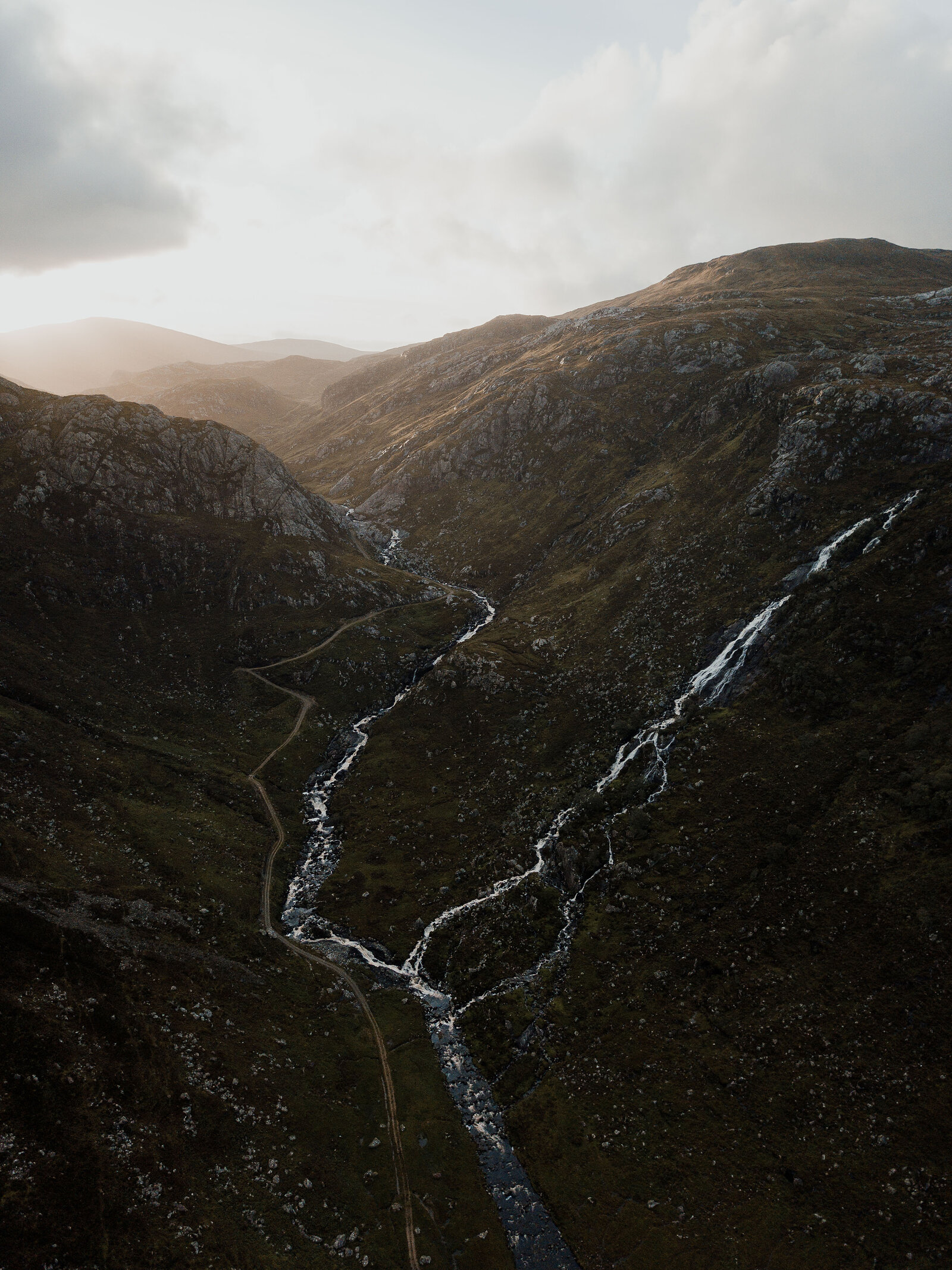 An aerial view of a mountain glen at dusk, a river forking into waterfalls between steep slopes.