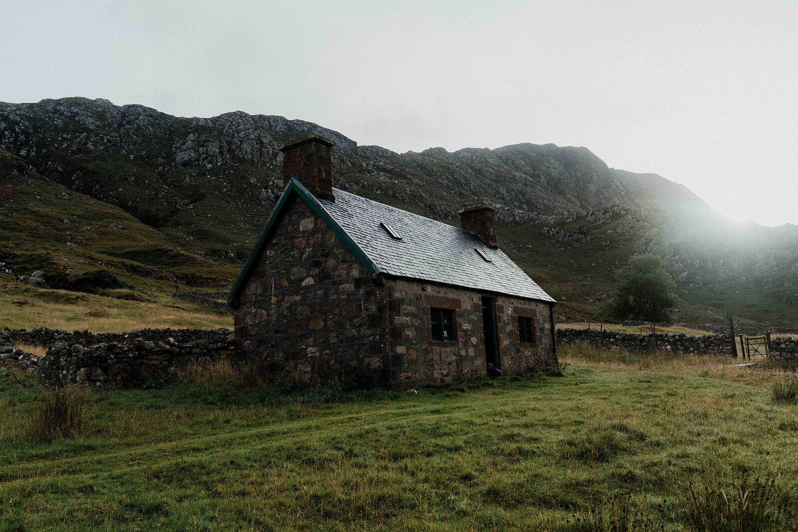 A stone cottage standing alone in a misty glen, mountains rising steeply behind its slate roof.