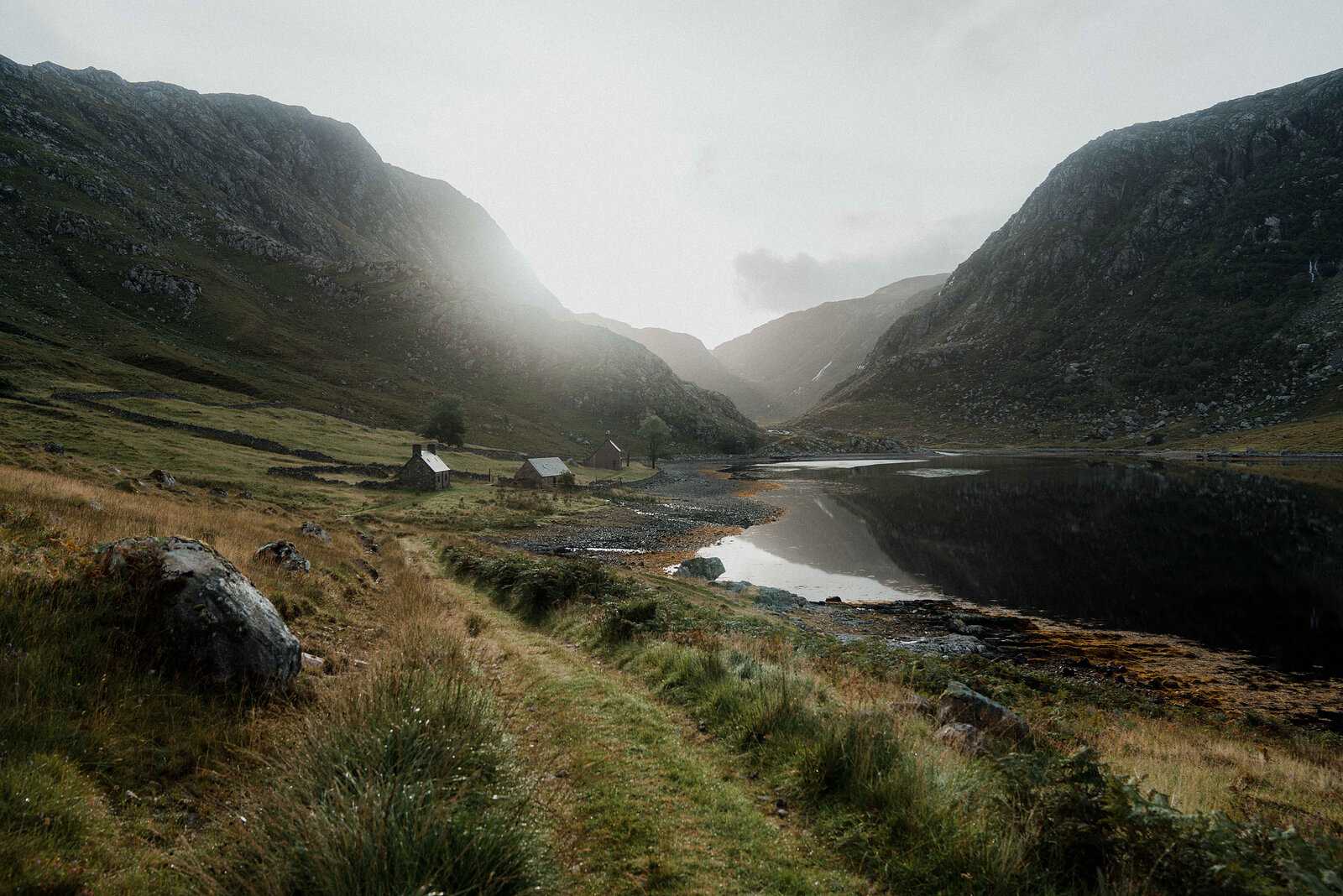 A cluster of stone cottages beside a loch in a misty mountain glen, a grassy path leading toward them.