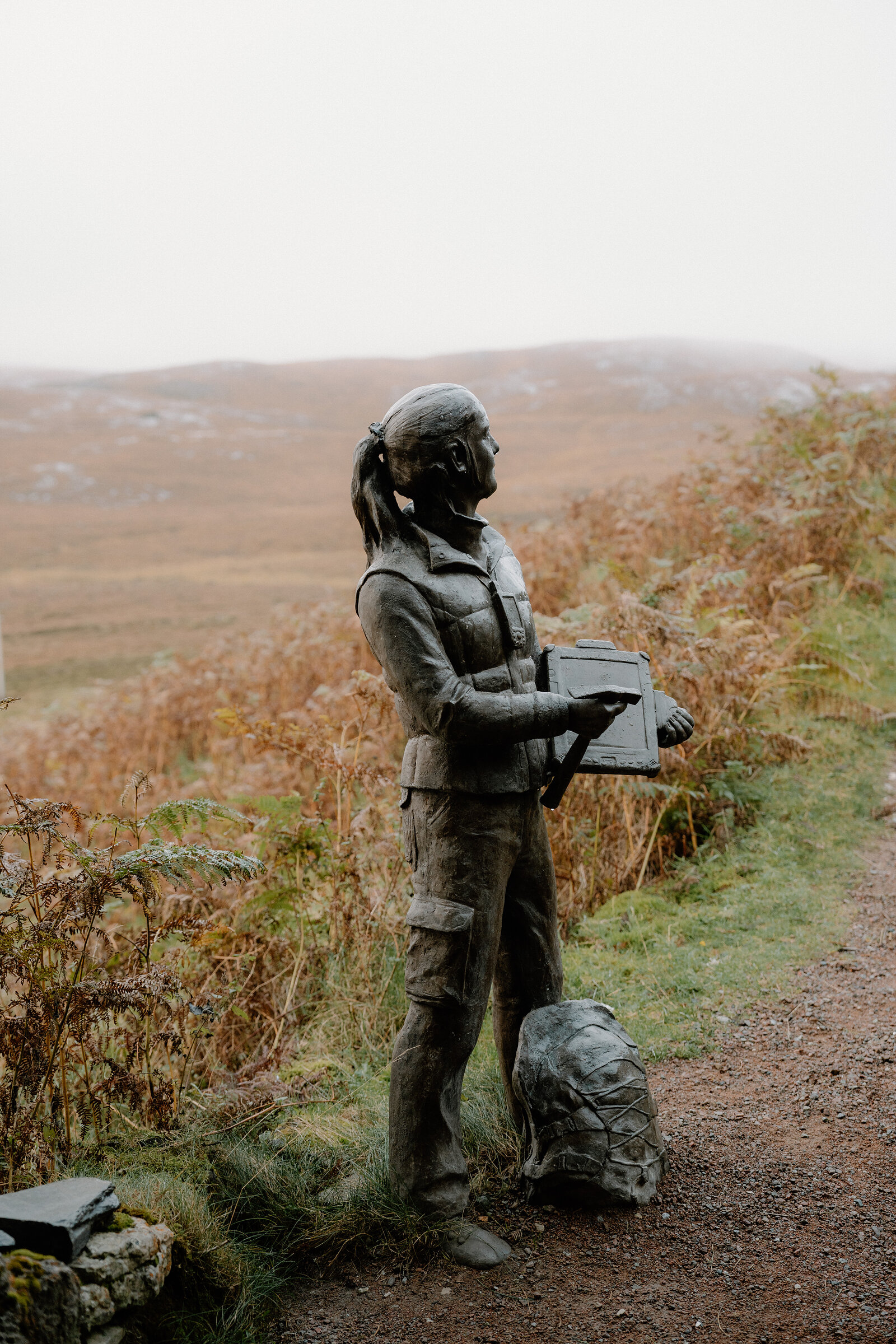 A bronze statue of a hiker with a camera and rucksack stands beside a gravel path on a misty moor.