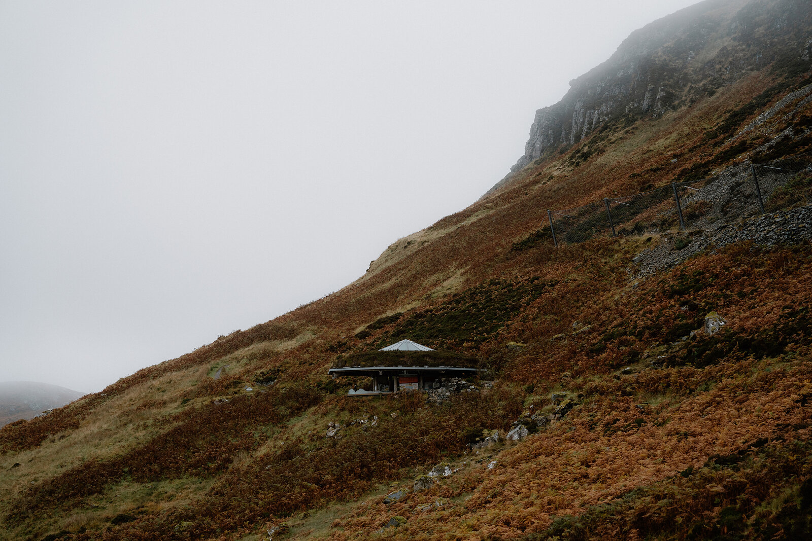 A small timber shelter sits on a heather-covered hillside shrouded in mist, its pitched roof just visible against the slope.