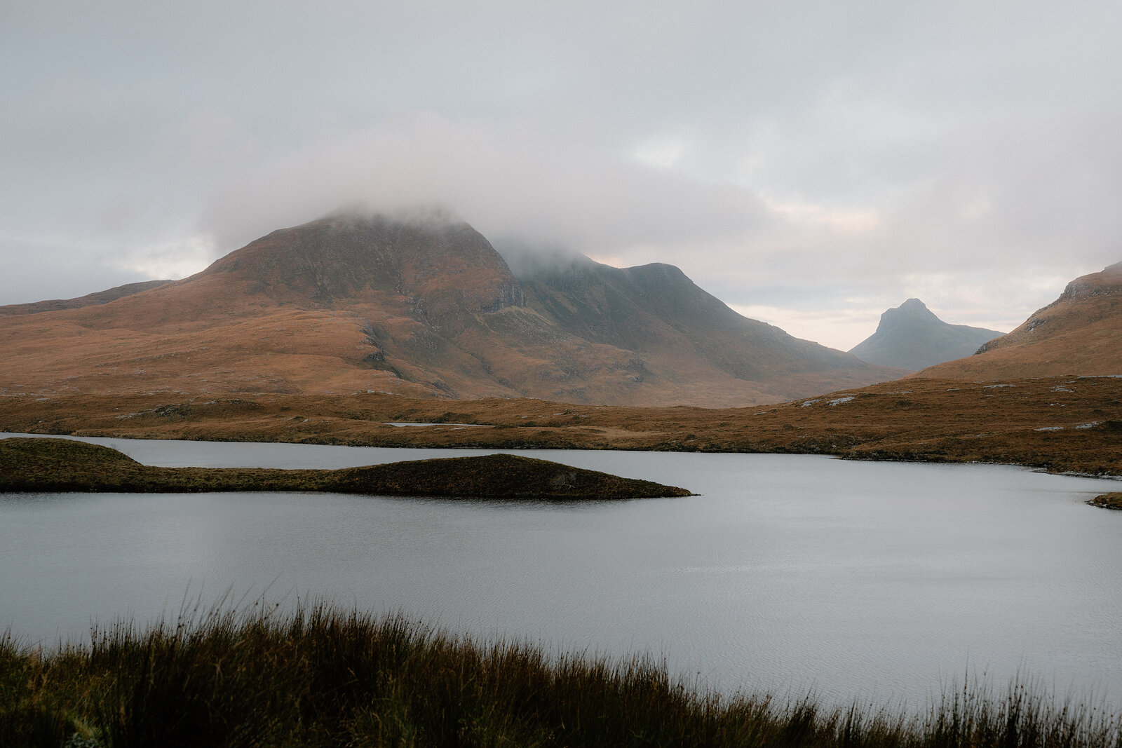 Mist wreathes a cluster of mountain peaks above a calm loch, heather moorland stretching into the foreground.