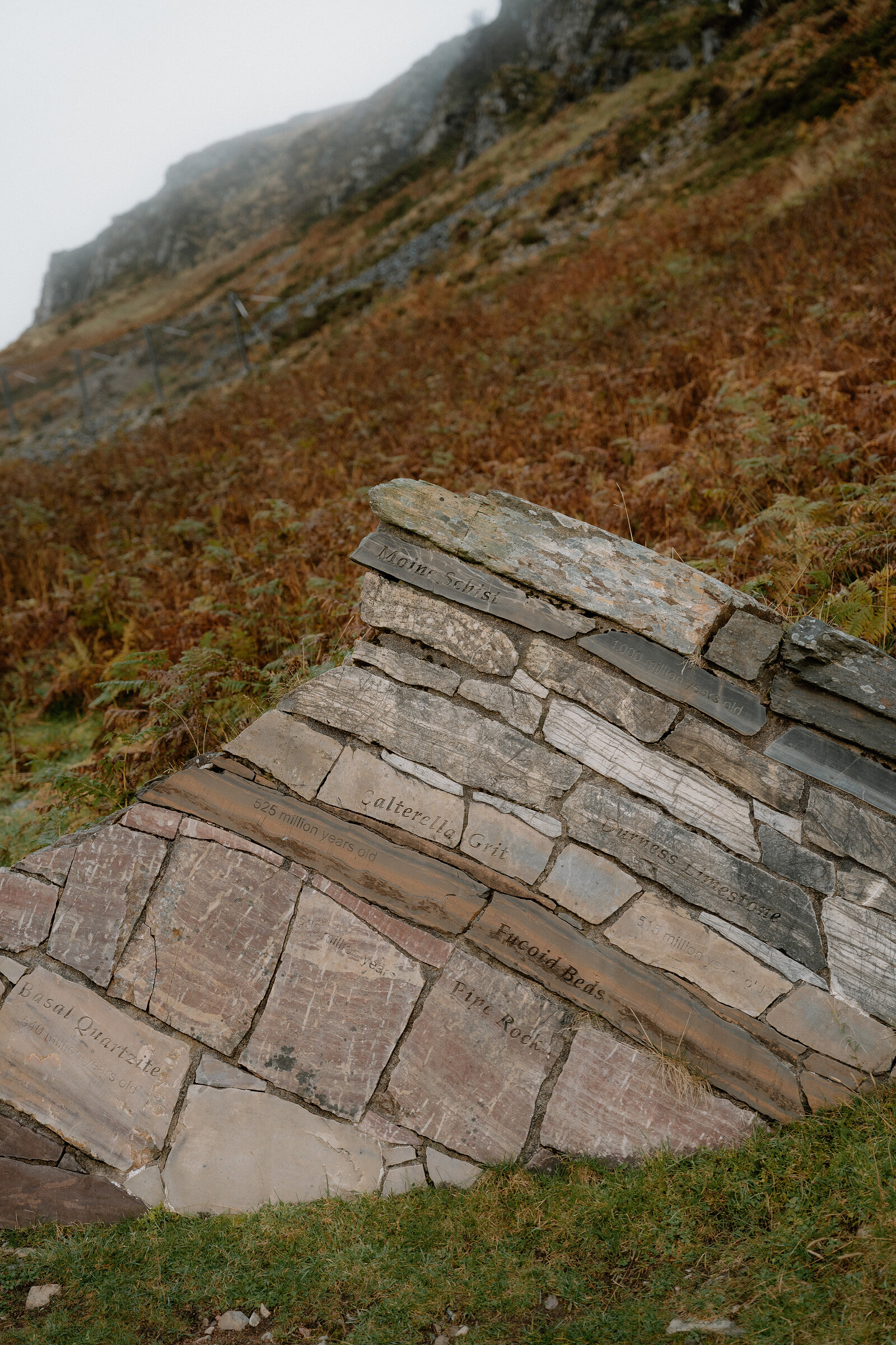 A stone wall carved with rock-type names like 'Wood Beds' and 'Fine Rock' stands on a misty hillside, part of a geological trail marker.