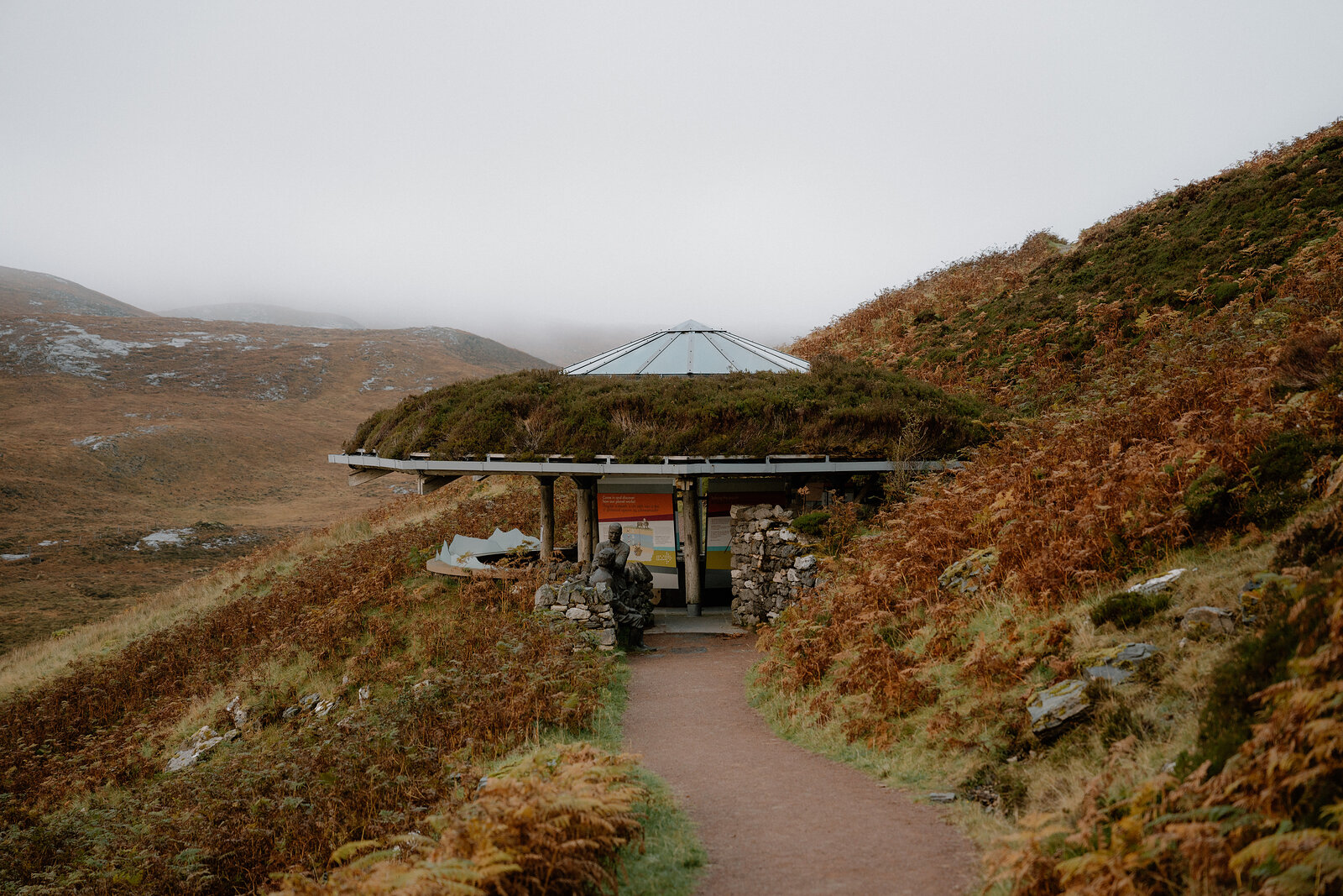 A circular wooden shelter with a domed roof sits beside a gravel path on a misty moor, low hills fading into fog beyond.