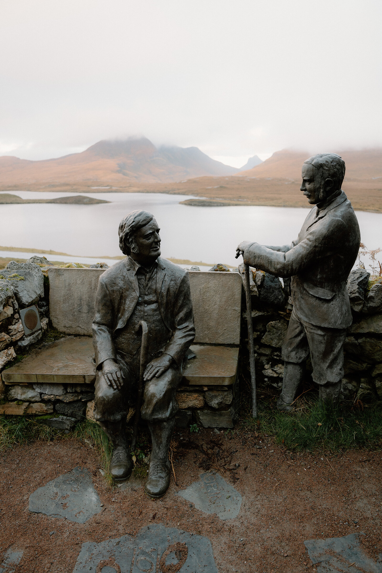Two bronze statues sit and stand on a stone bench overlooking a misty loch and distant mountains.