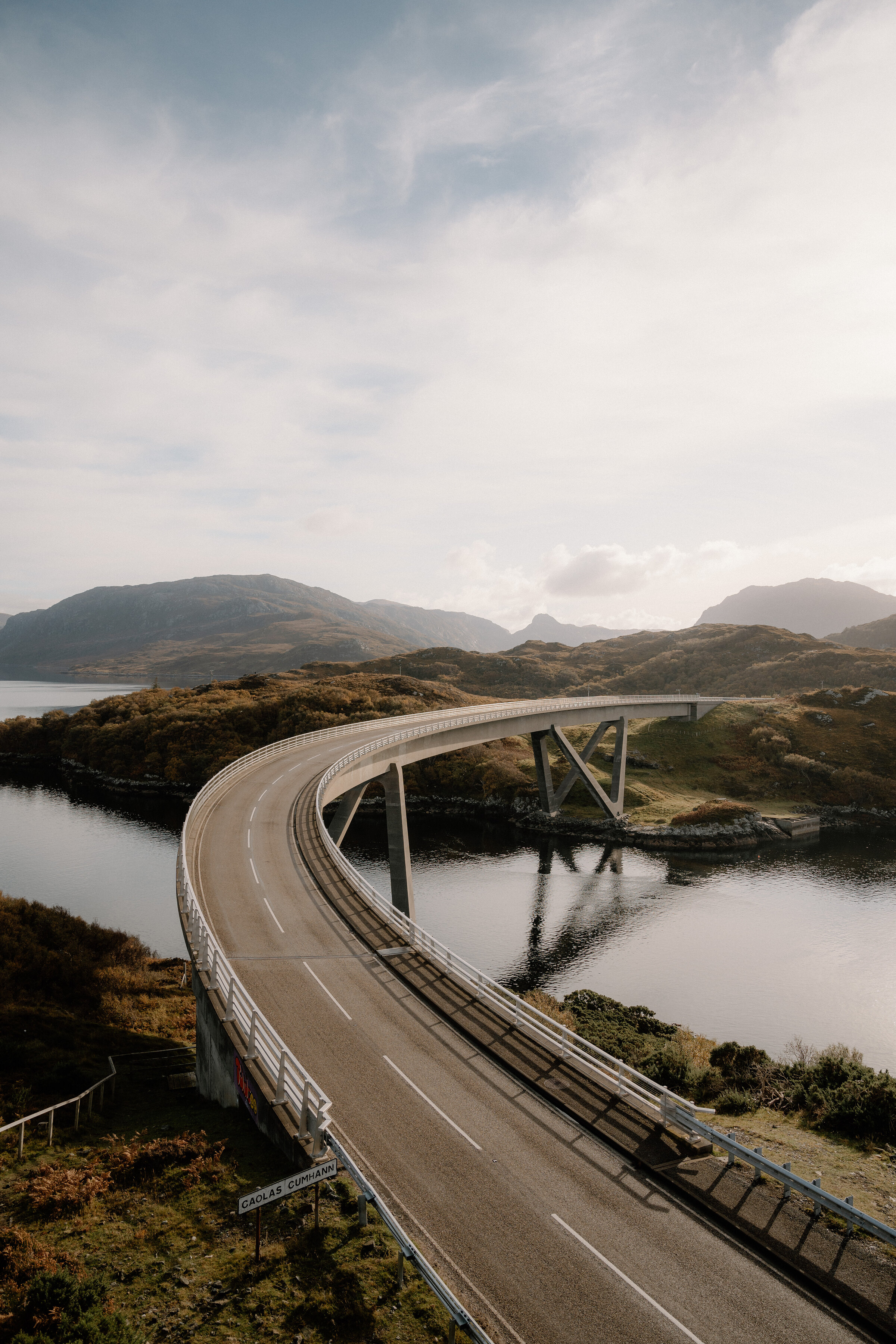 A curving road bridge sweeps over a sea loch between rocky hills, its white railings tracing the road's path across the water.