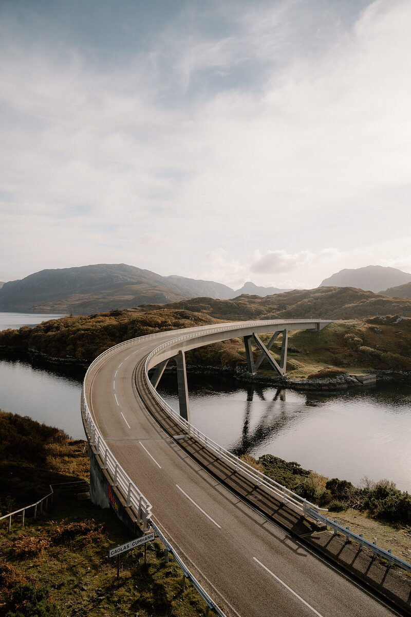 A curving road bridge sweeps over a sea loch between rocky hills, its white railings tracing the road's path across the water.