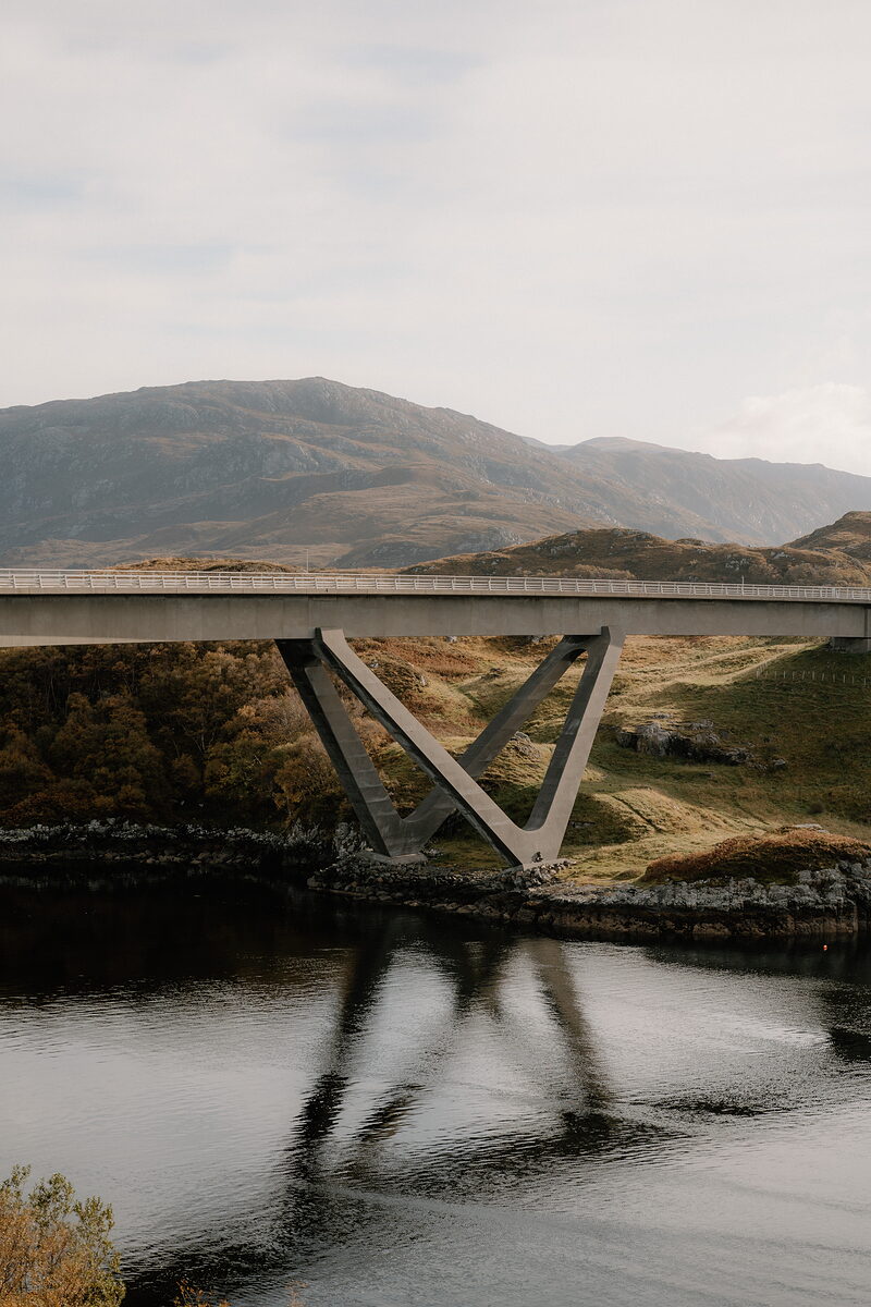 A road bridge with a V-shaped concrete support crosses a calm loch, mountains rising behind and the structure mirrored in the still water.