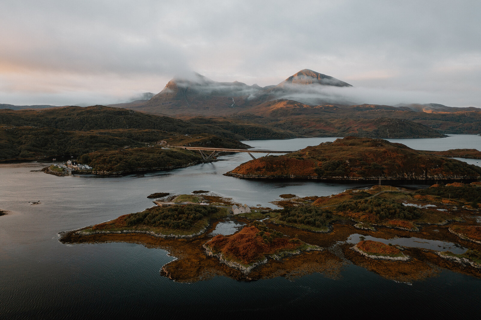 Layered rocky islets and inlets spread across a calm loch beneath mist-covered mountains, seen from an elevated angle.