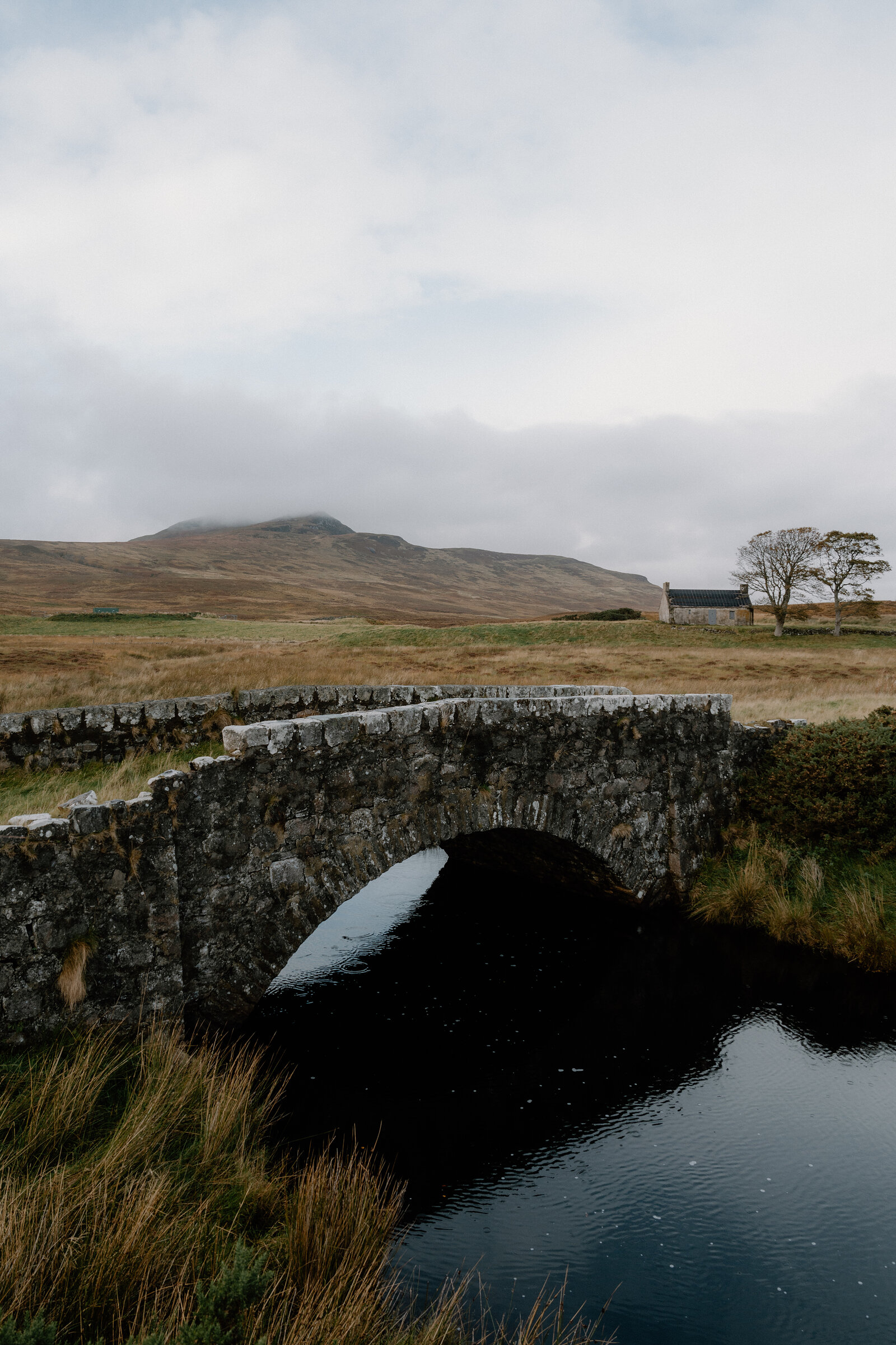 An old stone bridge arching over a stream, hills and a cottage in the distance.