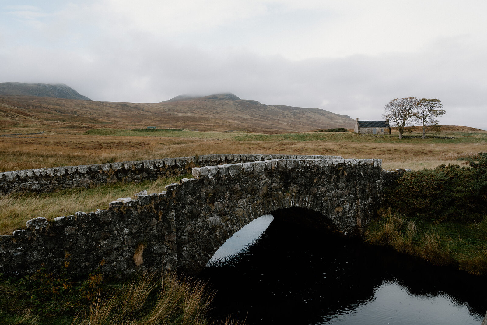 An old stone bridge arching over a stream, hills and a distant cottage beyond.