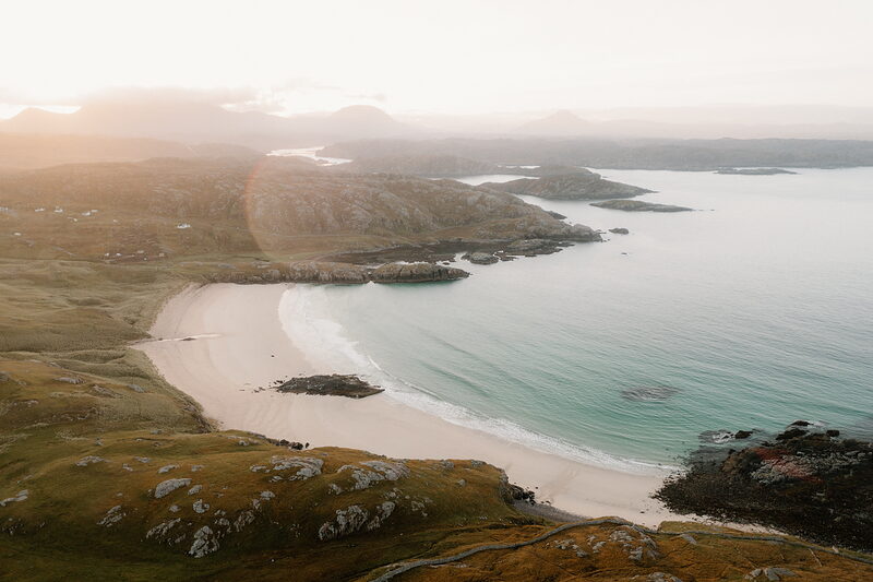 A sandy bay curves beneath golden evening light, hills rising along the coastline.