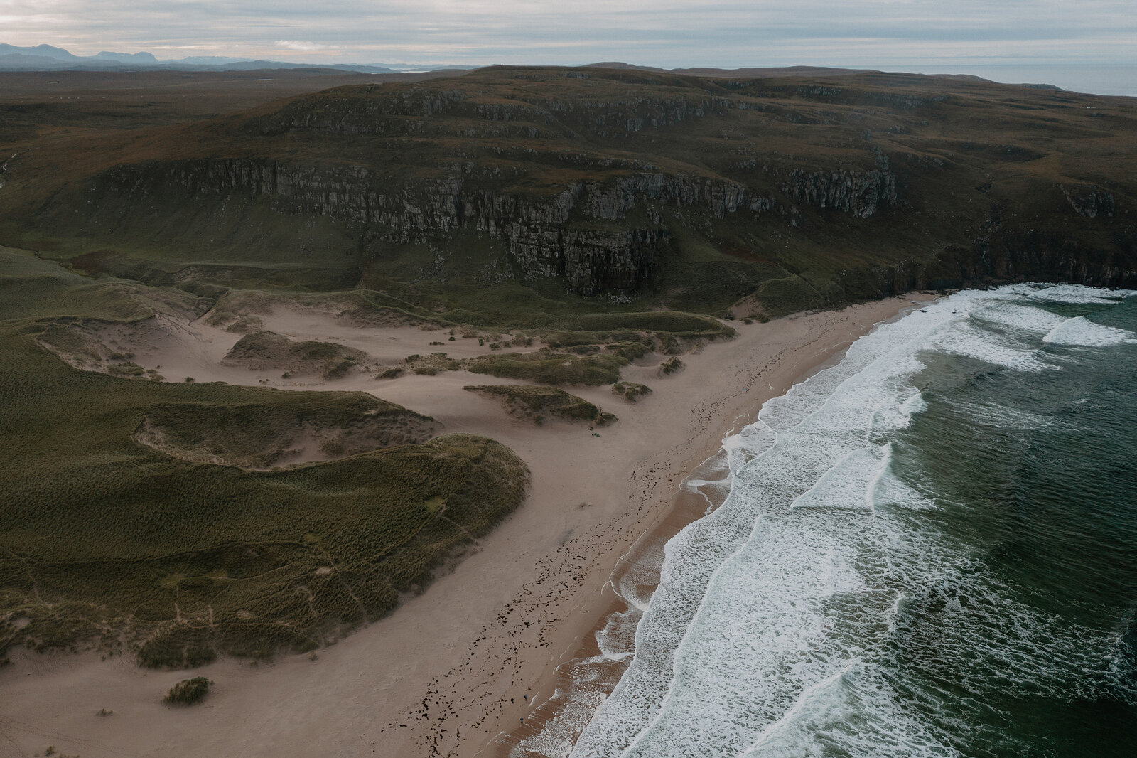 A sandy bay curves beneath dark cliffs, seen from above.