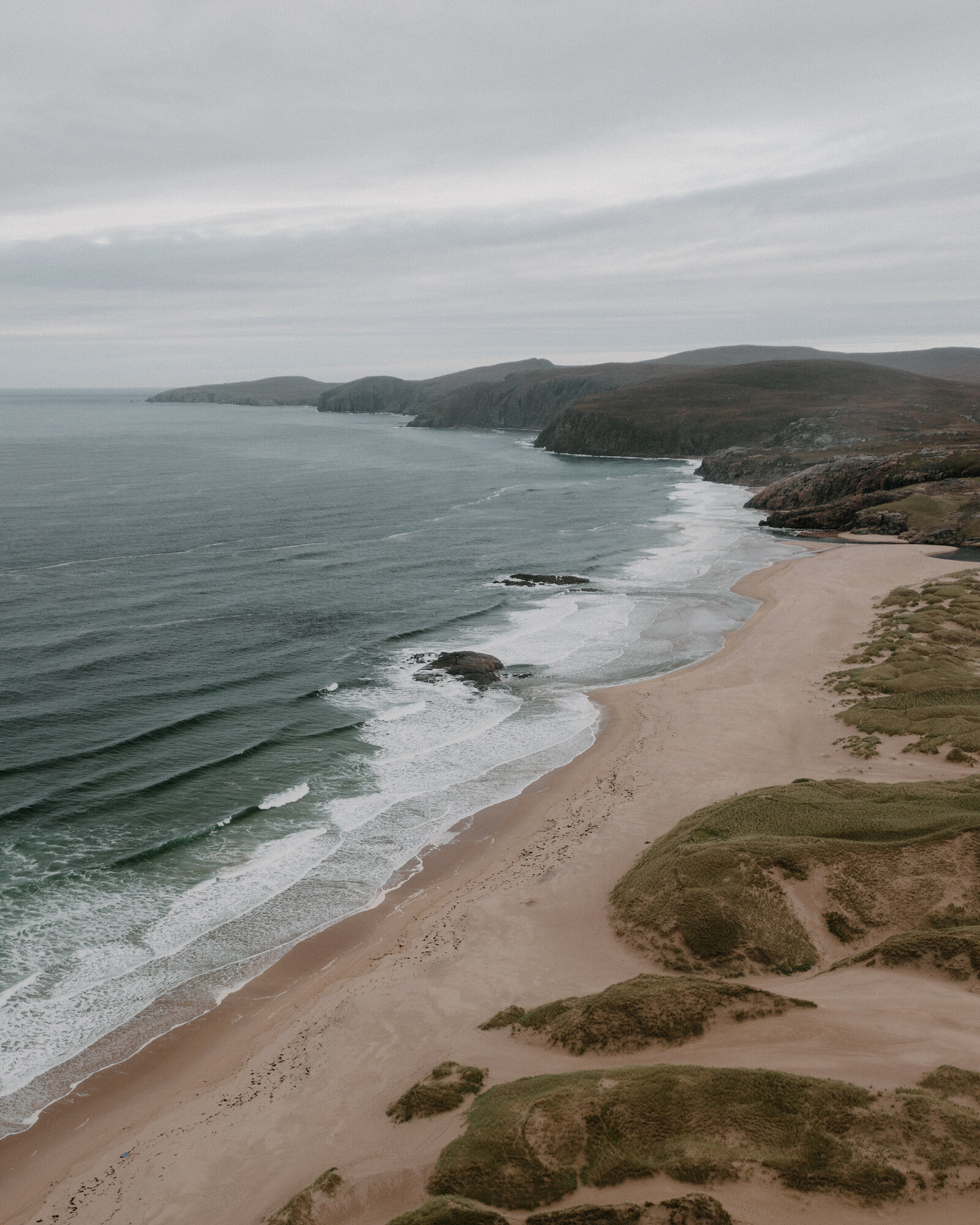 A sandy bay curves beneath rolling hills along the coastline, seen from above.