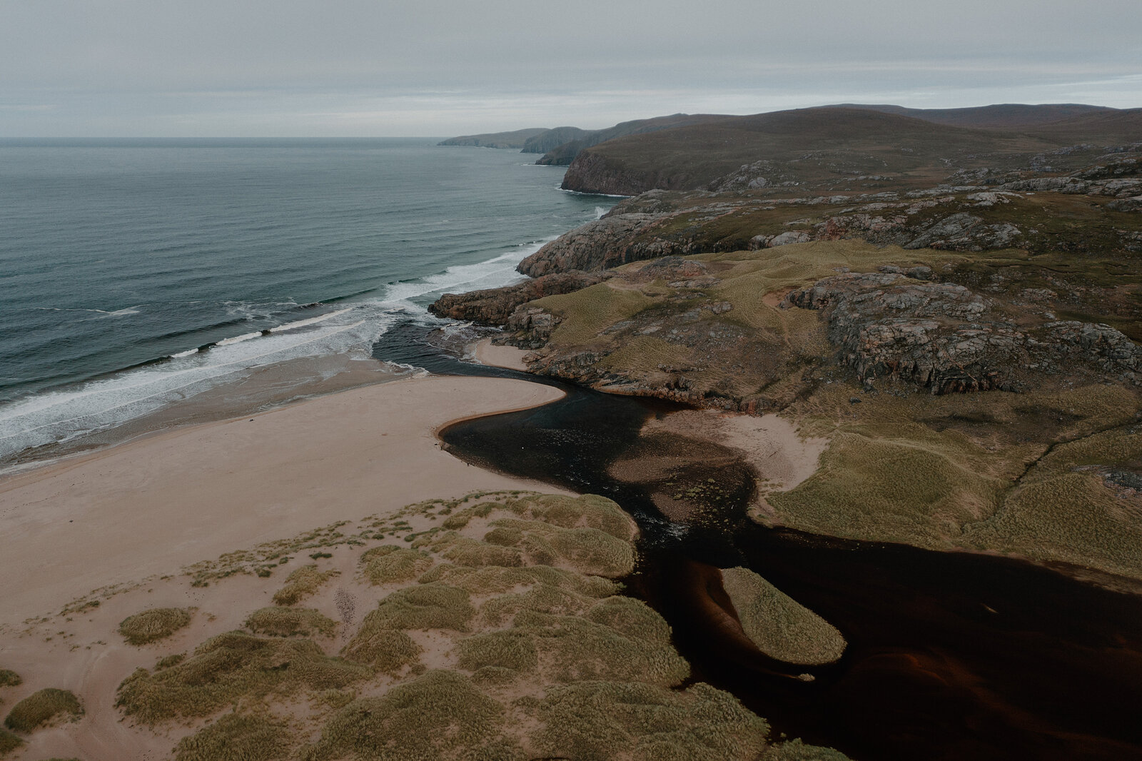 A dark peaty river winds across a sandy beach toward the sea, seen from above.