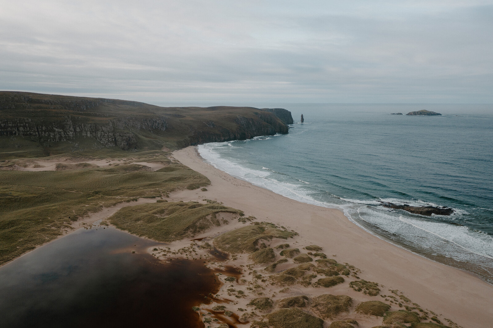 A rocky sea stack and small island sit off a sandy beach beneath grey skies.