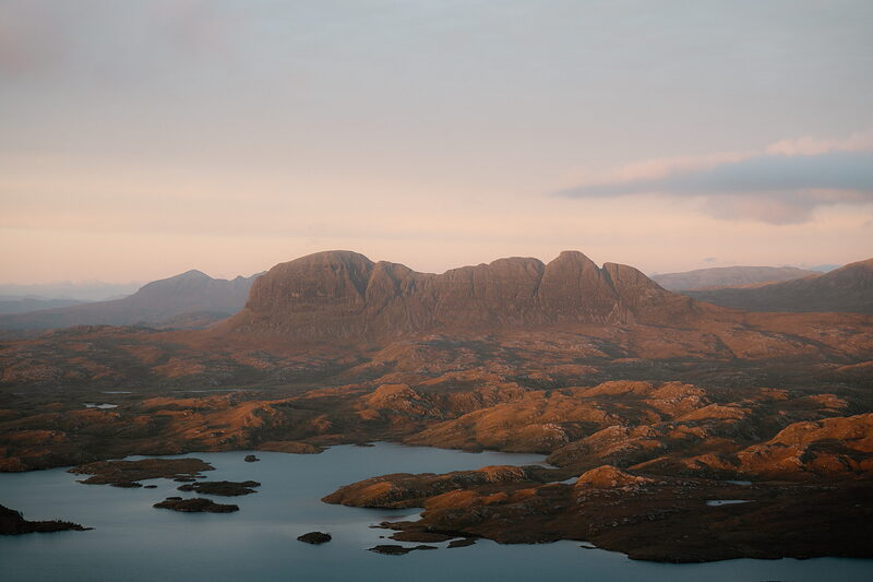 A rugged mountain peak catches the last light above a loch-dotted landscape.