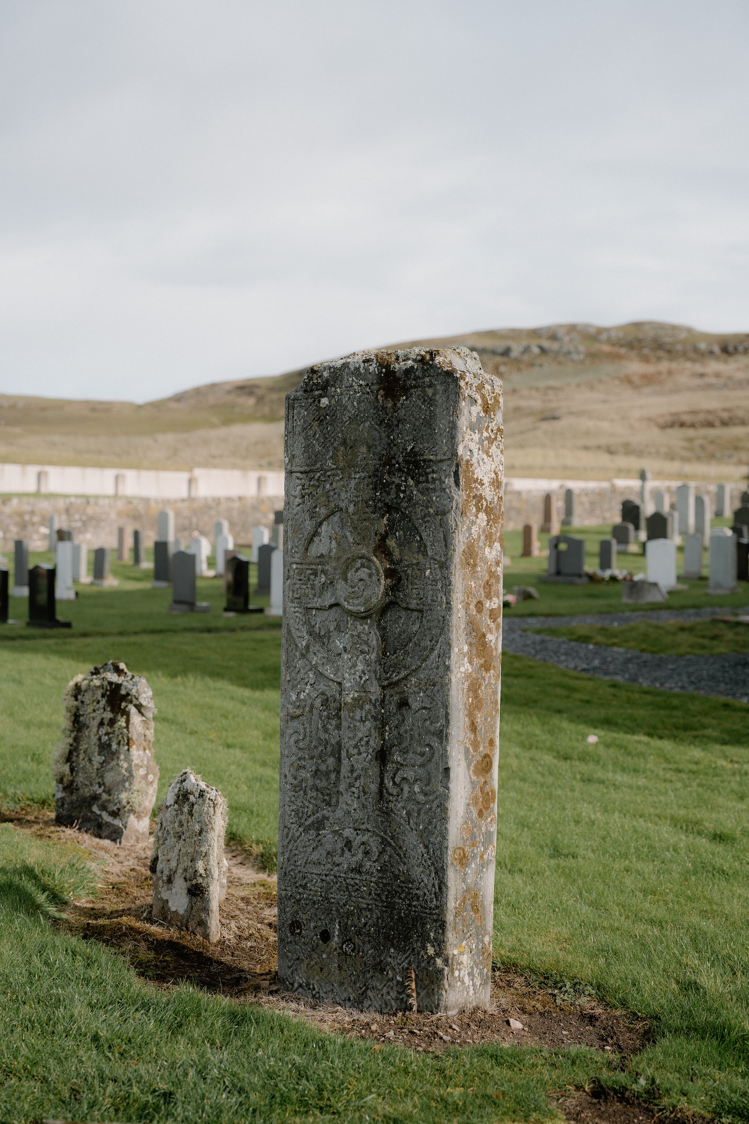 A carved Pictish cross-slab stands among gravestones in a coastal graveyard.