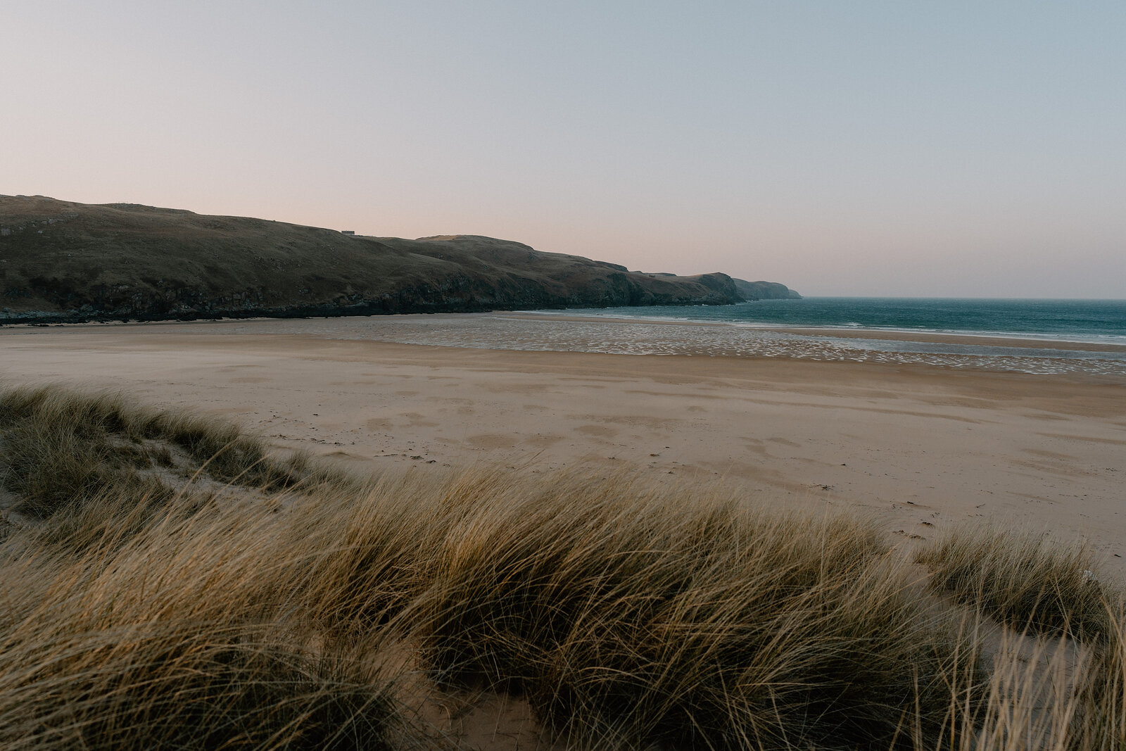 Dune grass frames a wide sandy beach beneath hills at dusk.