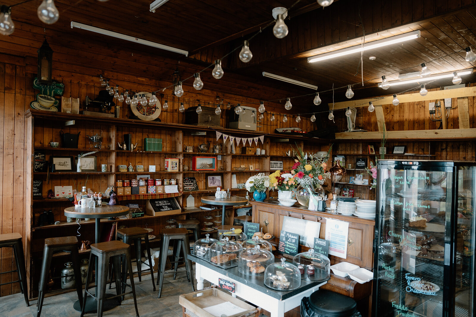 A glass-domed cake display sits on a counter inside a rustic timber-panelled café.