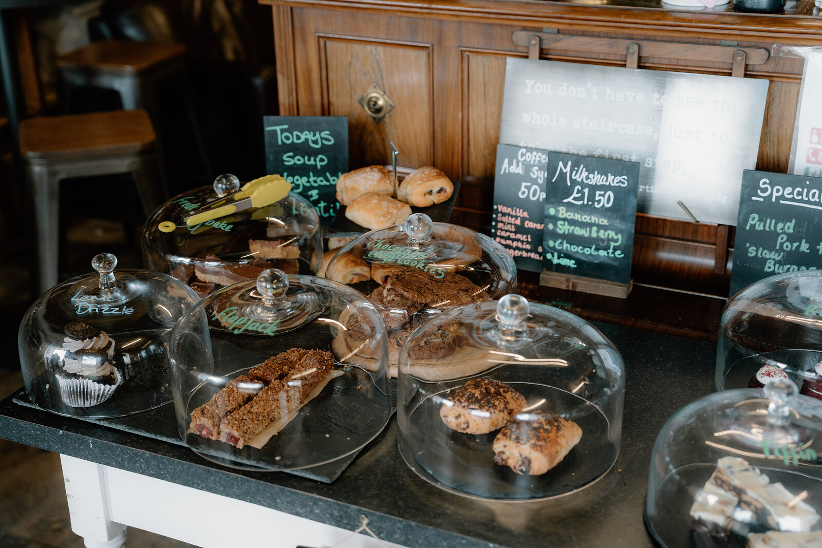 Cakes and pastries sit beneath glass domes on a counter inside a rustic timber-panelled café.