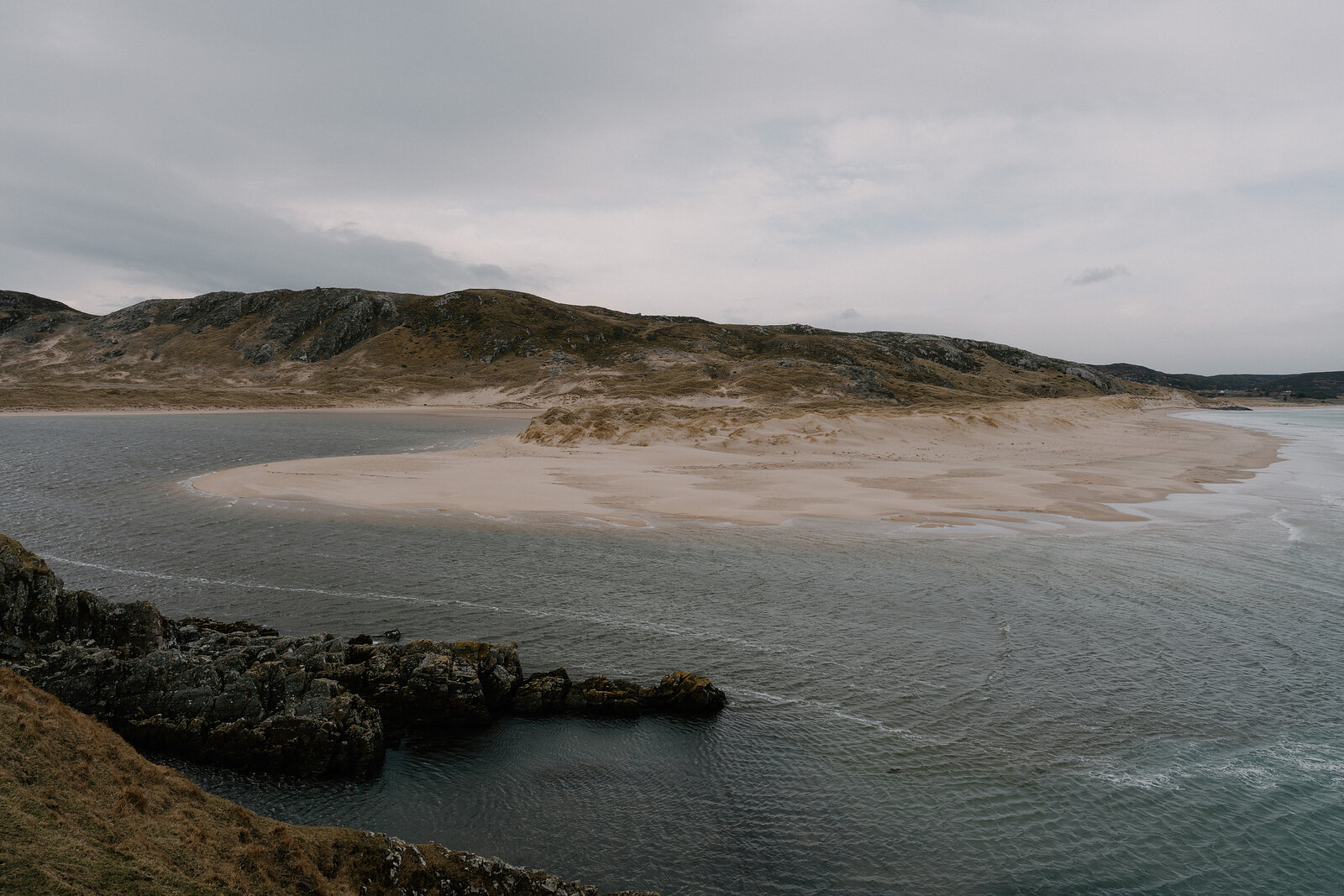 A sandy tidal spit curves out into a sea inlet below rounded, rocky hills.