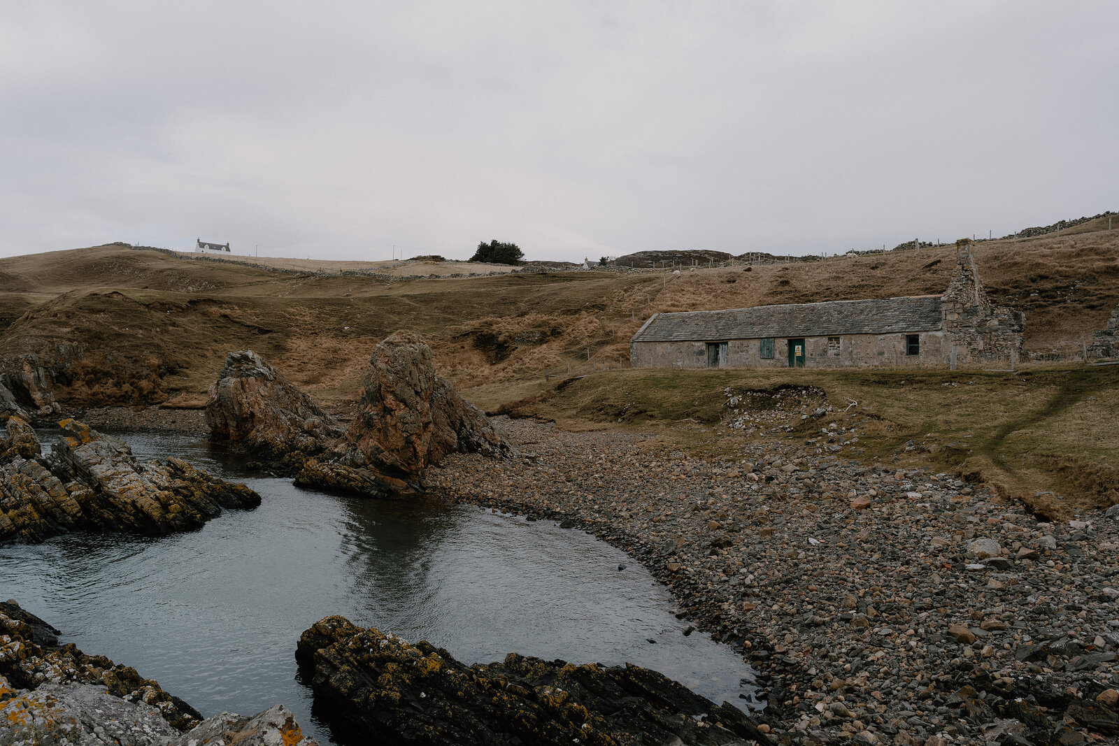 A ruined stone cottage stands beside a rocky shore.
