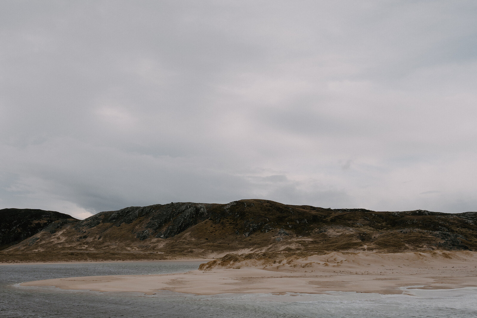 A sandy beach curves beneath a dune-covered headland under an overcast sky.