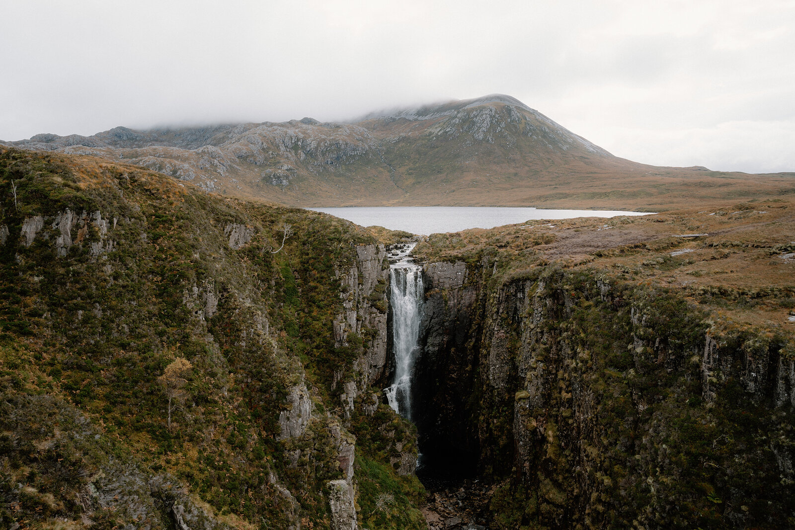 A waterfall drops through a narrow gorge below a still loch, with a mountain rising behind.