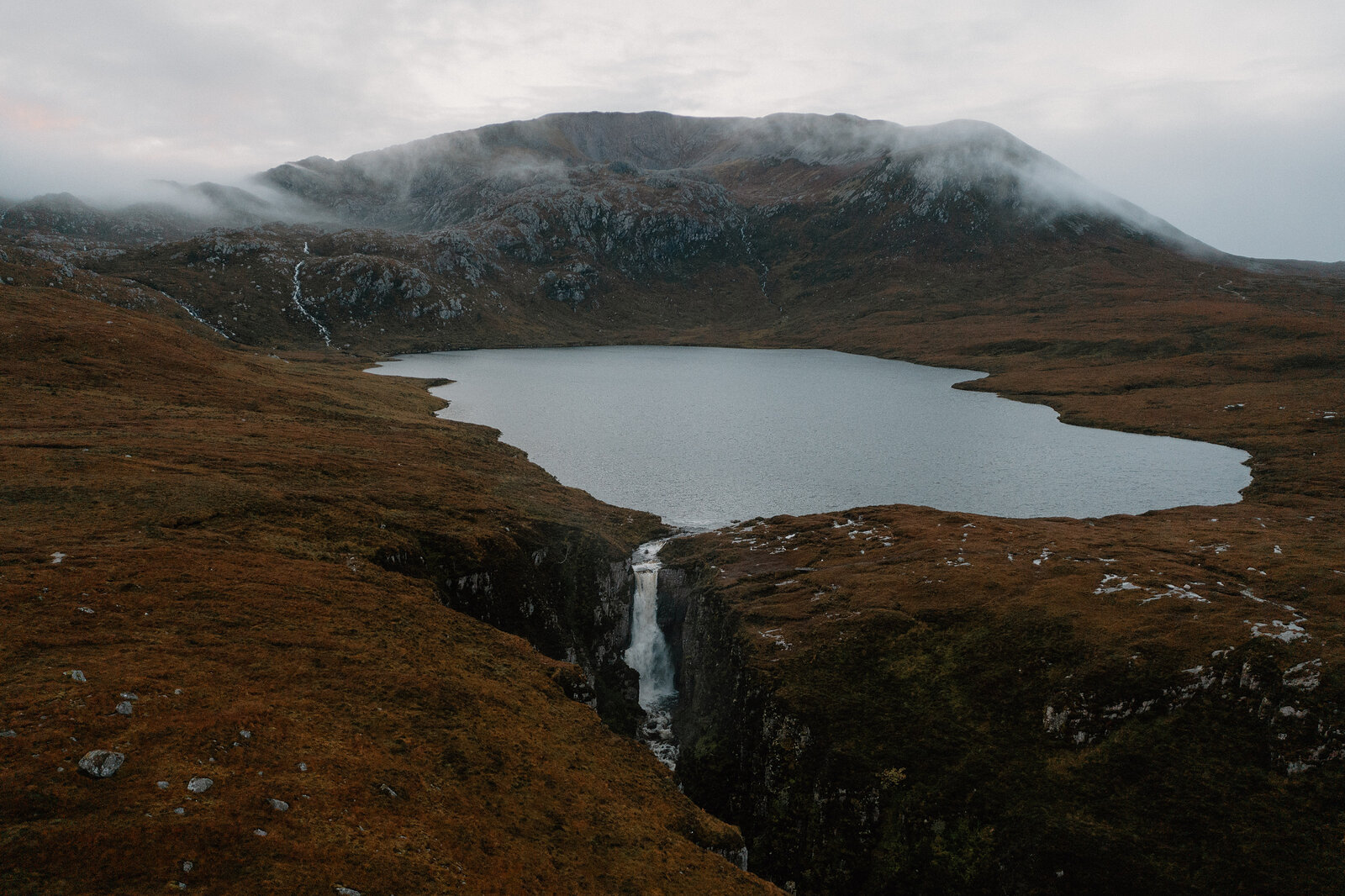 A loch sits below a cloud-wrapped mountain, water spilling from it into a waterfall at the gorge's edge.