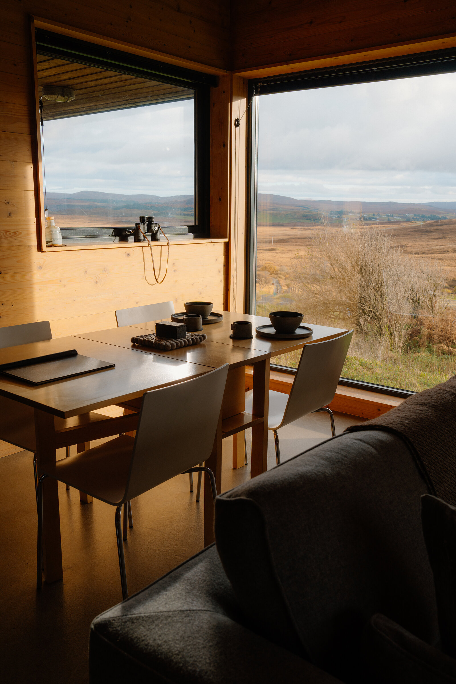 A wooden dining table set for two beside a corner window, with binoculars on the sill and a wide view of moorland hills beyond; a sofa arm in the foreground.