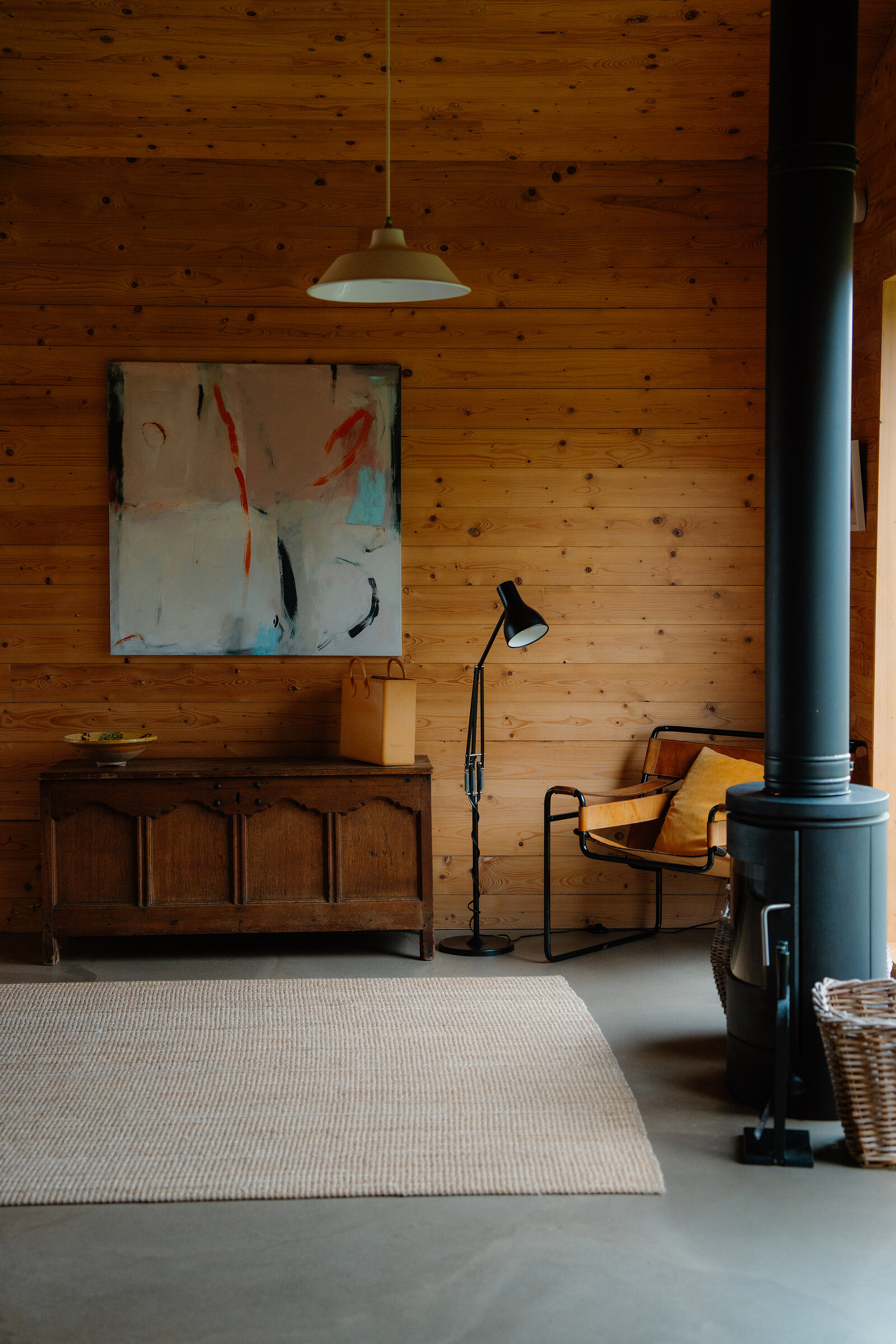 A timber-clad room with an abstract painting above an old panelled chest, a floor lamp and a chair with a striped cushion beside a black stove flue, on a polished concrete floor with a woven rug.