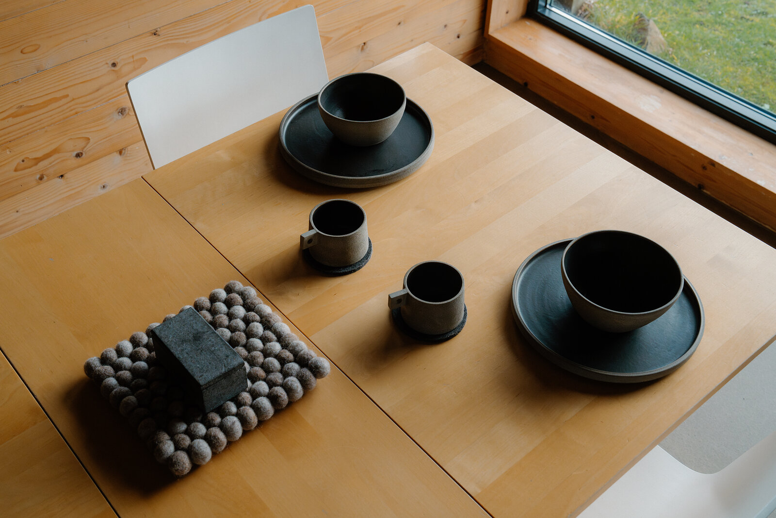 A set of dark grey ceramic tableware including bowls, cups and saucers, laid out on a wooden table beside a stack of felted pompom coasters and a plain chair, with a window looking onto grass beyond.