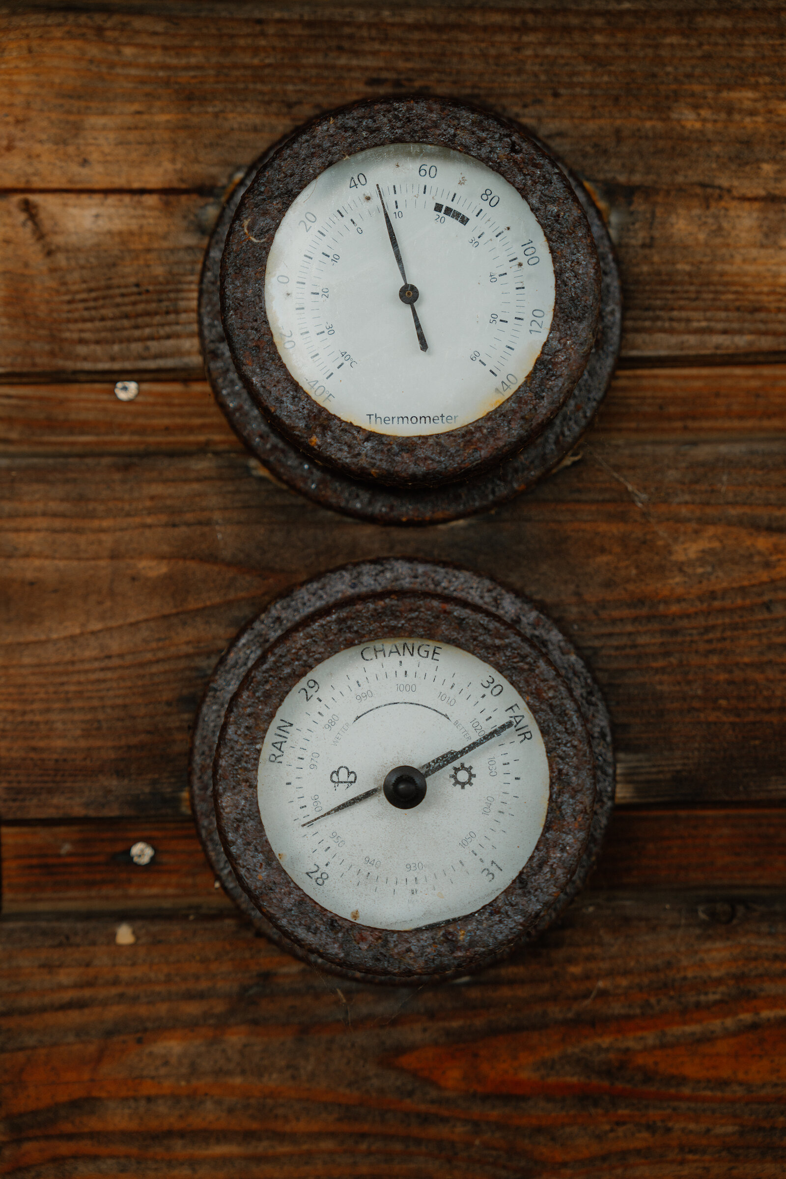 Two rust-edged weather gauges, a thermometer and a barometer marked rain to fair, mounted side by side on a wooden plank wall.