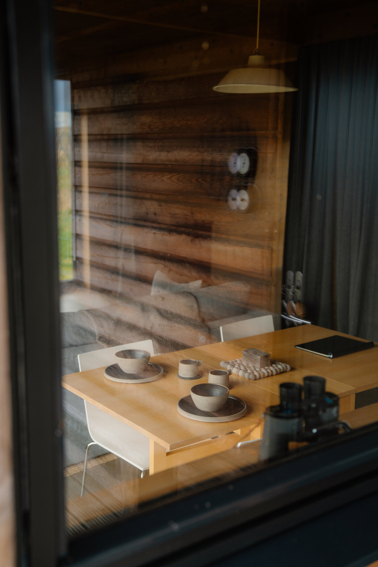 A wooden dining table set with dark ceramic bowls, cups and a bowl of eggs, seen through a window reflecting a sofa and pendant light inside the cabin.