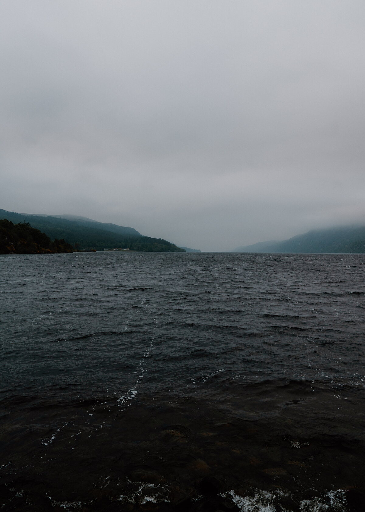A misty loch stretches between wooded hills, the far shore fading into cloud.
