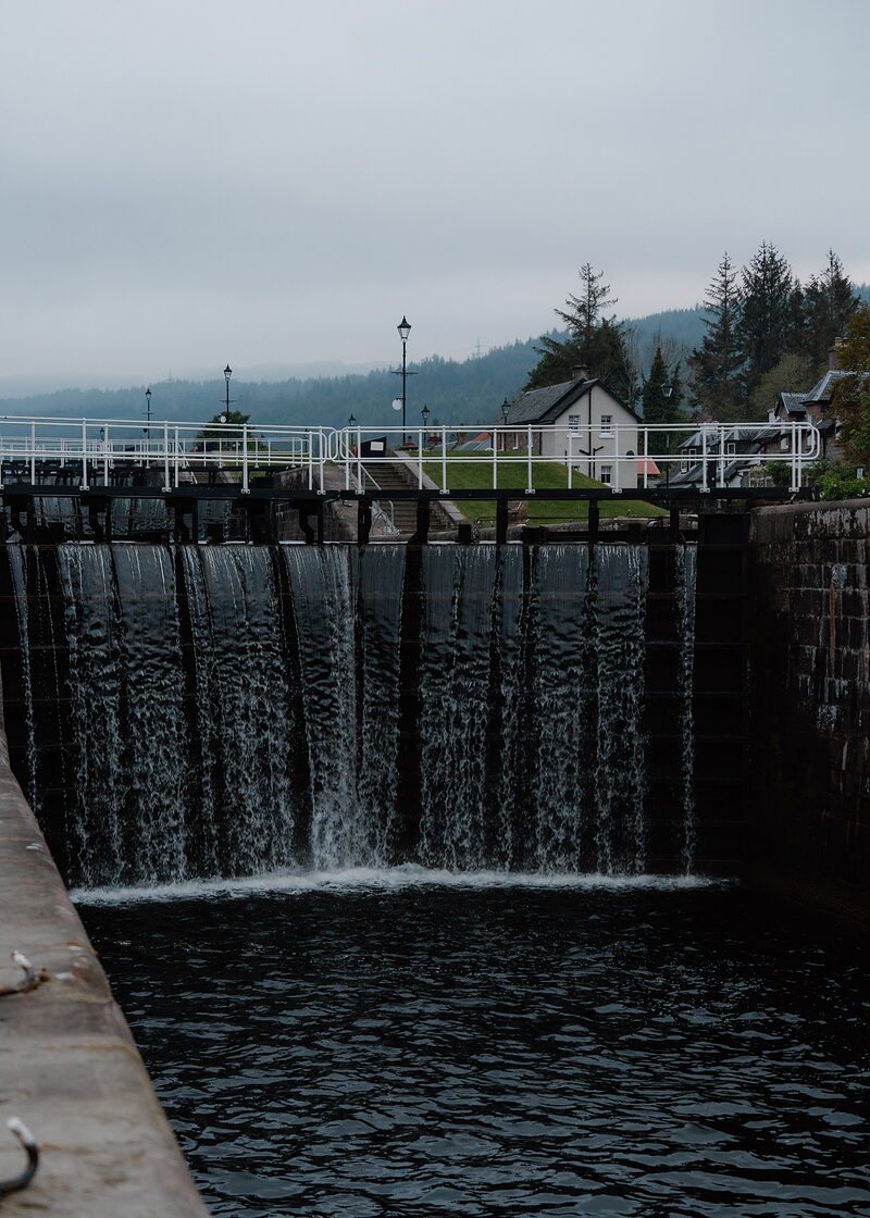 Water pours through open canal lock gates into a dark basin, white railings running along the lock walls and a lockkeeper's cottage and pine trees standing behind in mist.