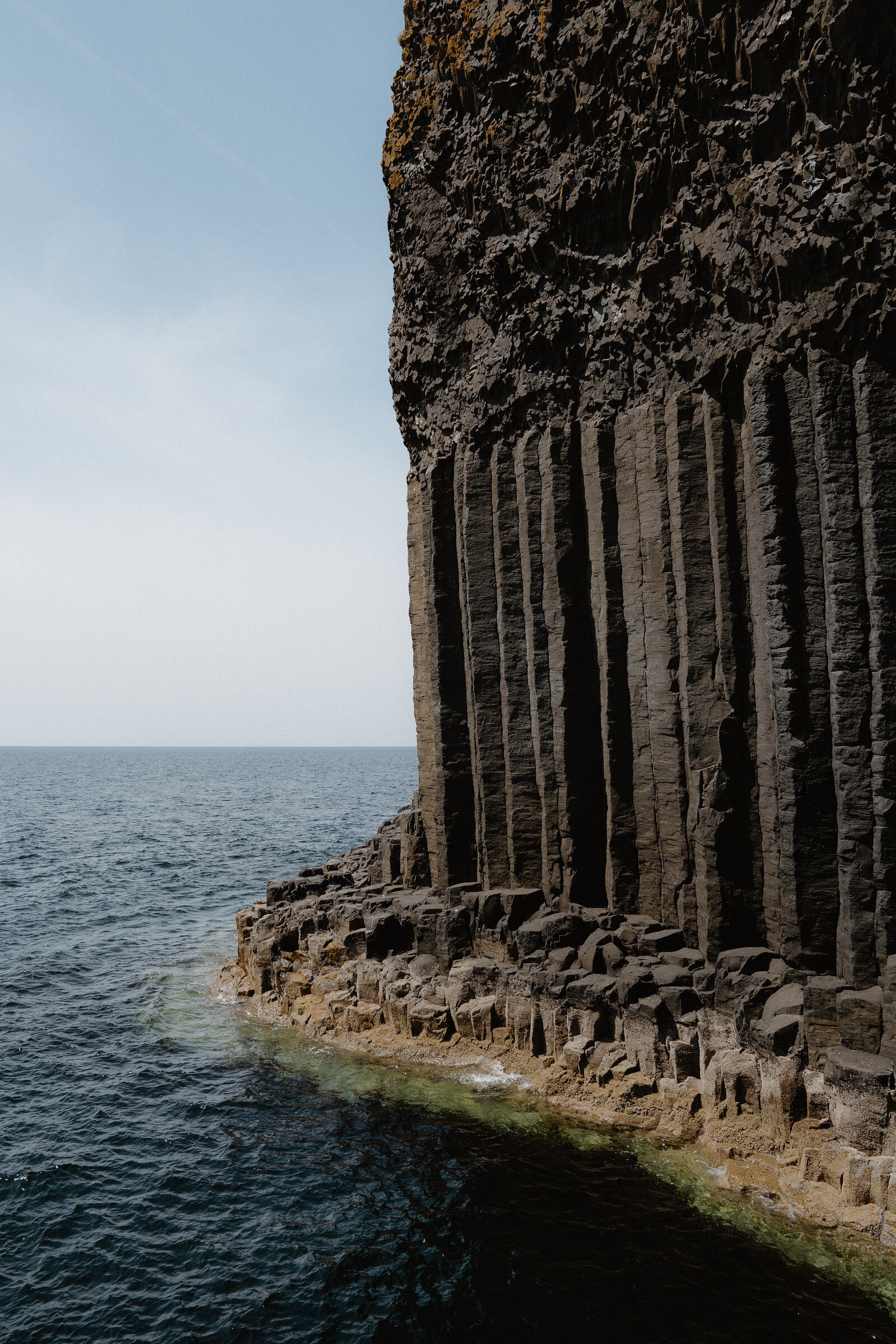 A cliff face of tightly packed hexagonal basalt columns rising from the sea, pale rock and shallow water pooling at its base.