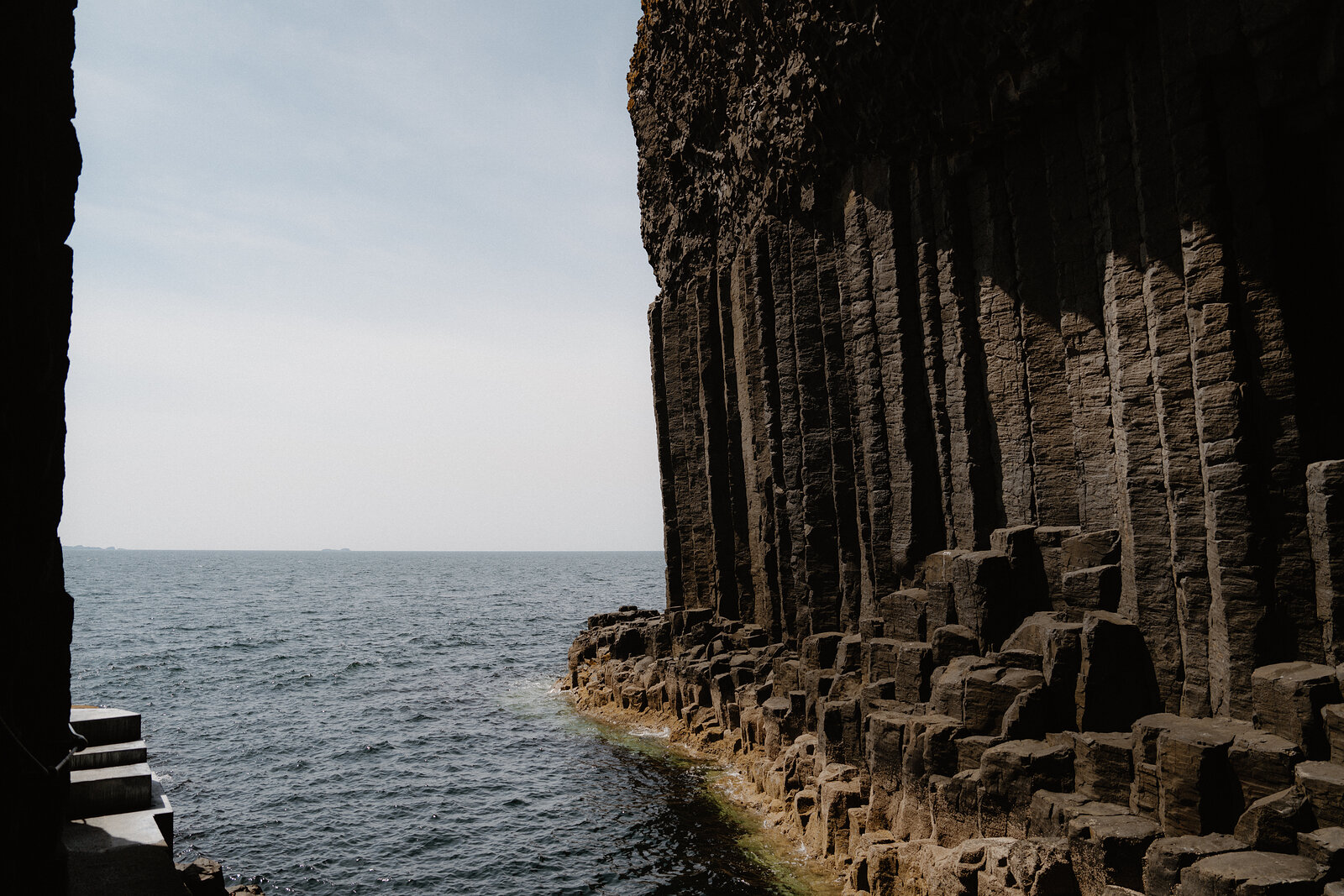Rows of dark basalt columns rise beside open water, a distant boat a small speck on the horizon.