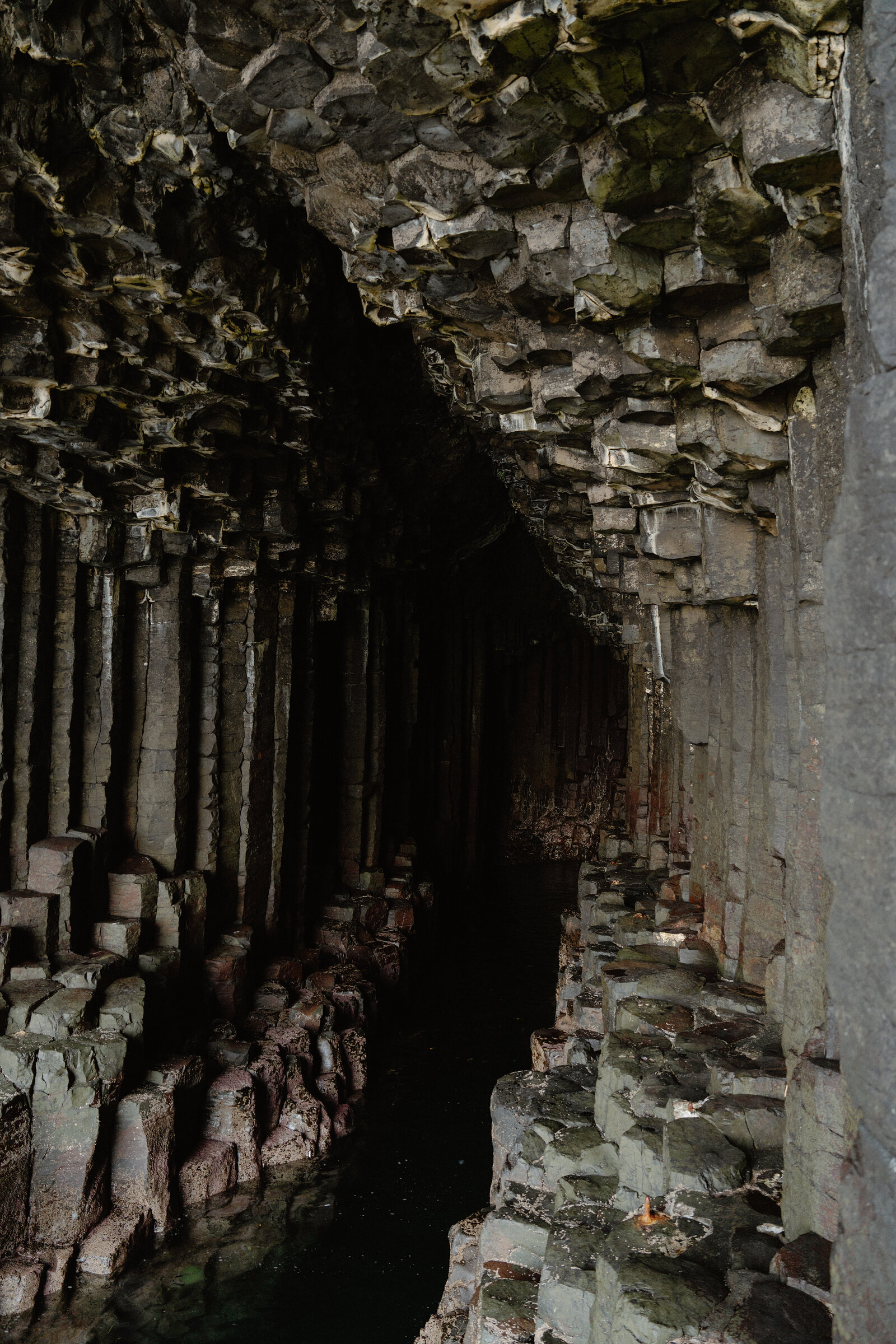 A narrow passage between basalt columns floods with still water, the rock reflected dark and green in its surface.