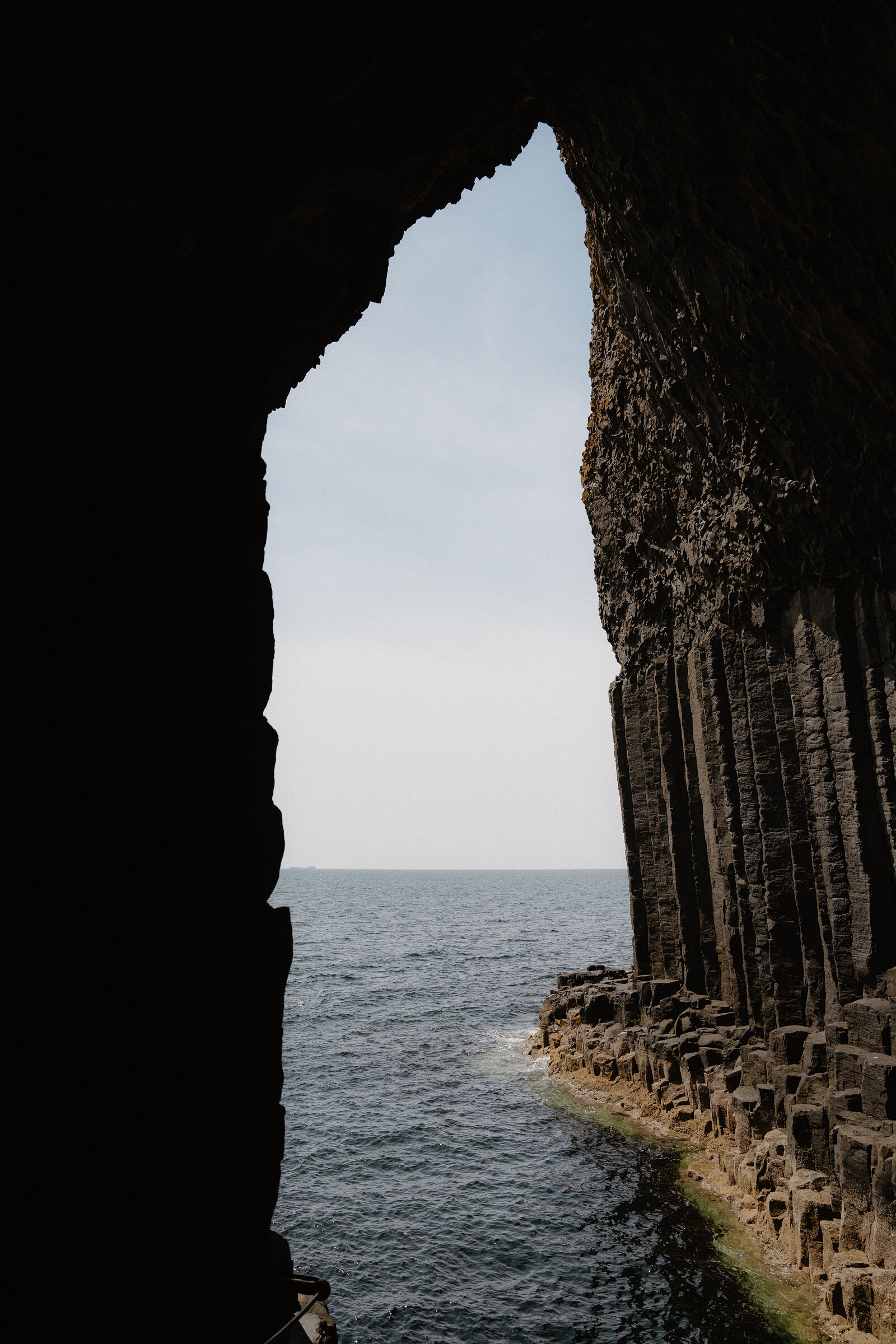 A dark basalt cave mouth frames a view of open sea, sunlit rock columns lining one side of the arched opening.
