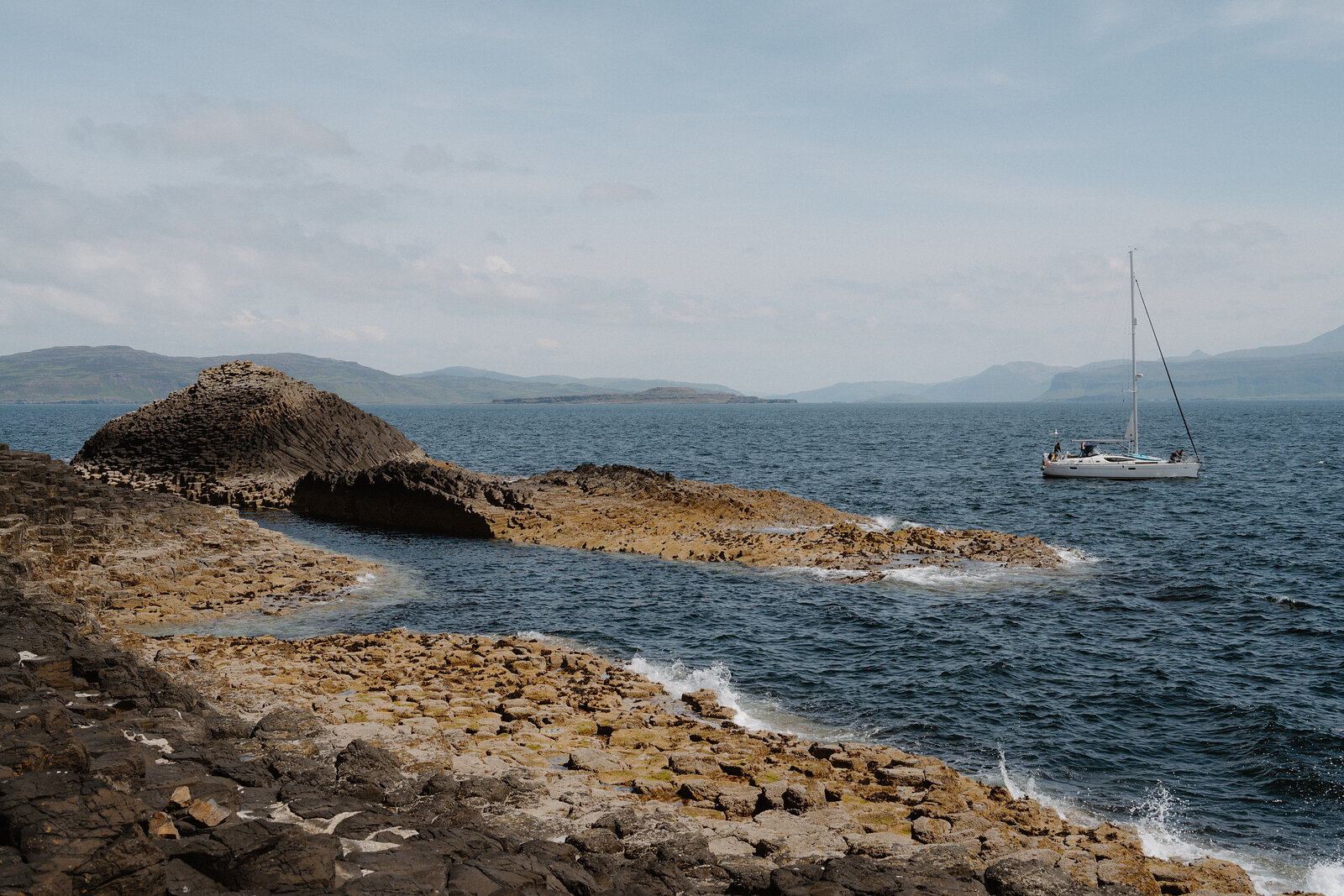 A low basalt rock arch rises at the shoreline, a sailing boat anchored beyond it and hills visible across the water.
