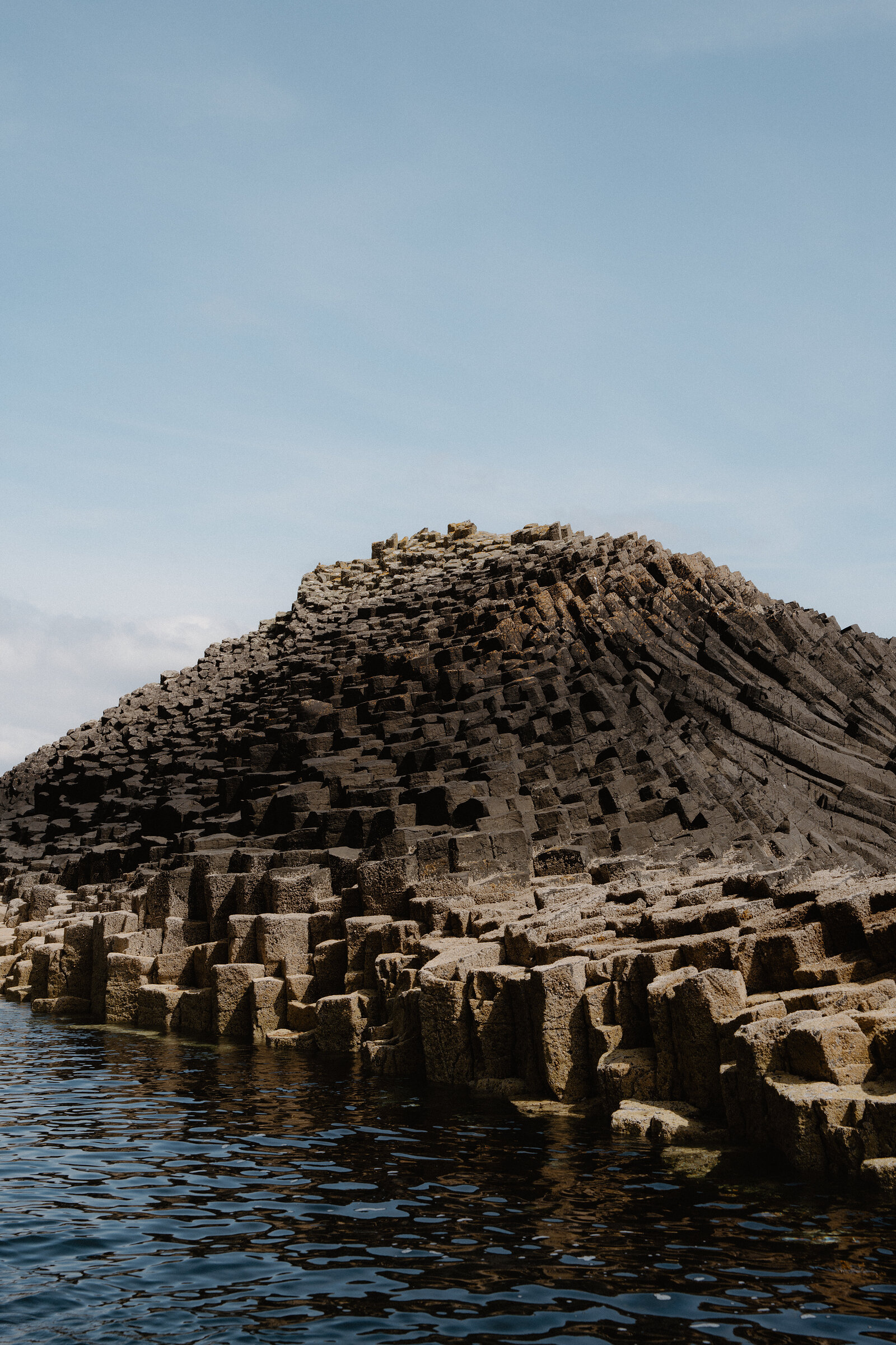 A conical mound of basalt columns rises steeply from the sea, water lapping at the exposed rock around its base.