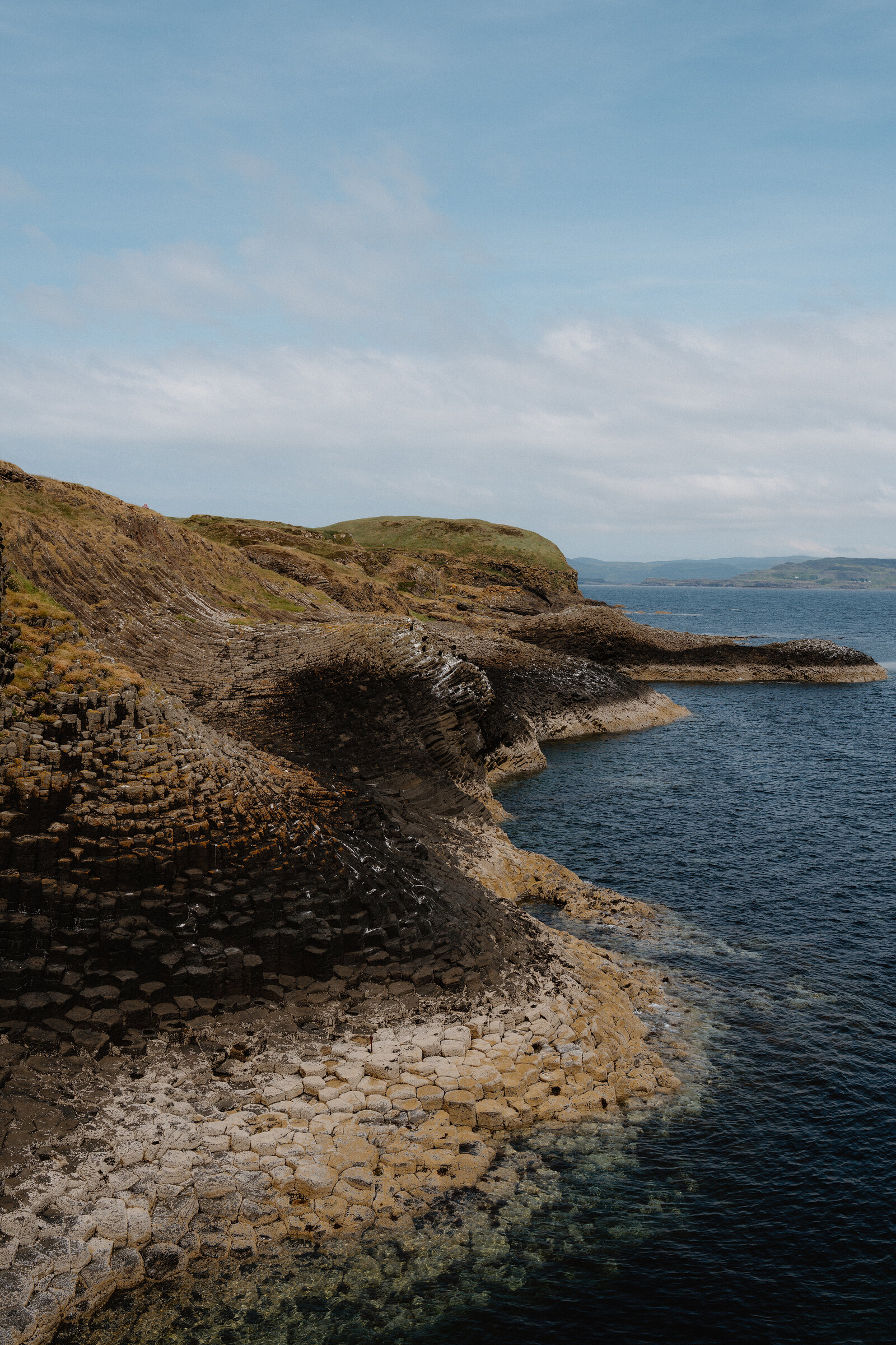 A headland of basalt columns steps down to the sea, distant hills visible across the water.