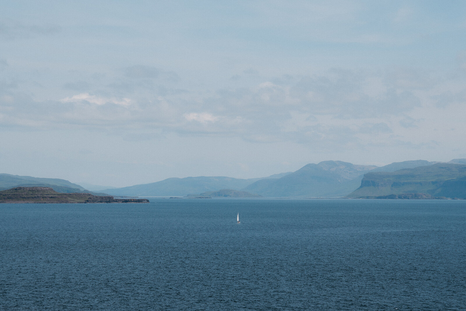 A small sailing boat crosses open water beneath layered hills fading into haze.